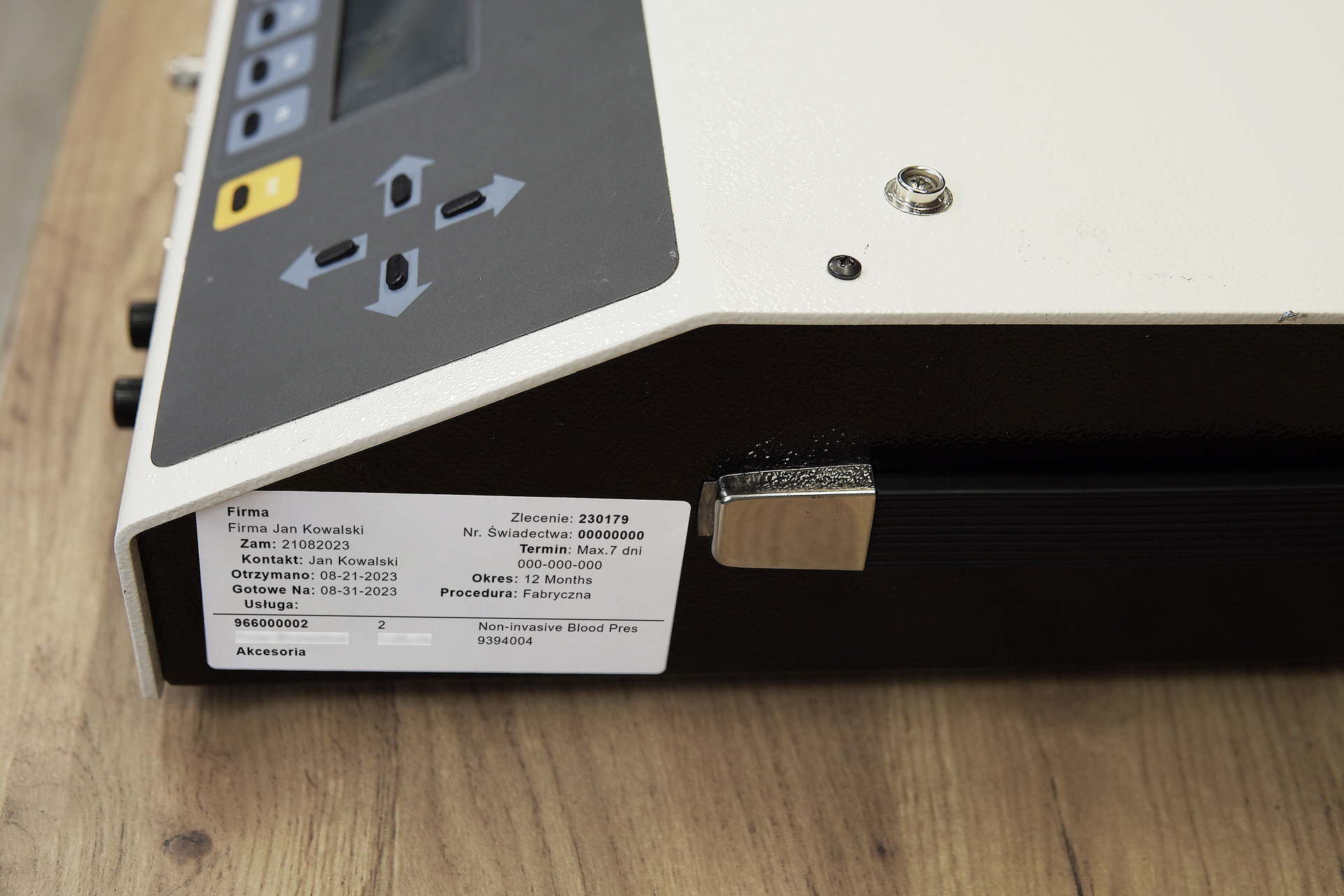 Zdjęcie przedstawia symulator nieinwazyjnego mierzenia ciśnienia tętniczego, w skrócie NIBP w perspektywie od boku. Na bocznej ścianie obudowy znajduje się naklejka z informacjami o urządzeniu. Widoczny jest częściowo panel sterujący z przyciskami oraz ekranem.