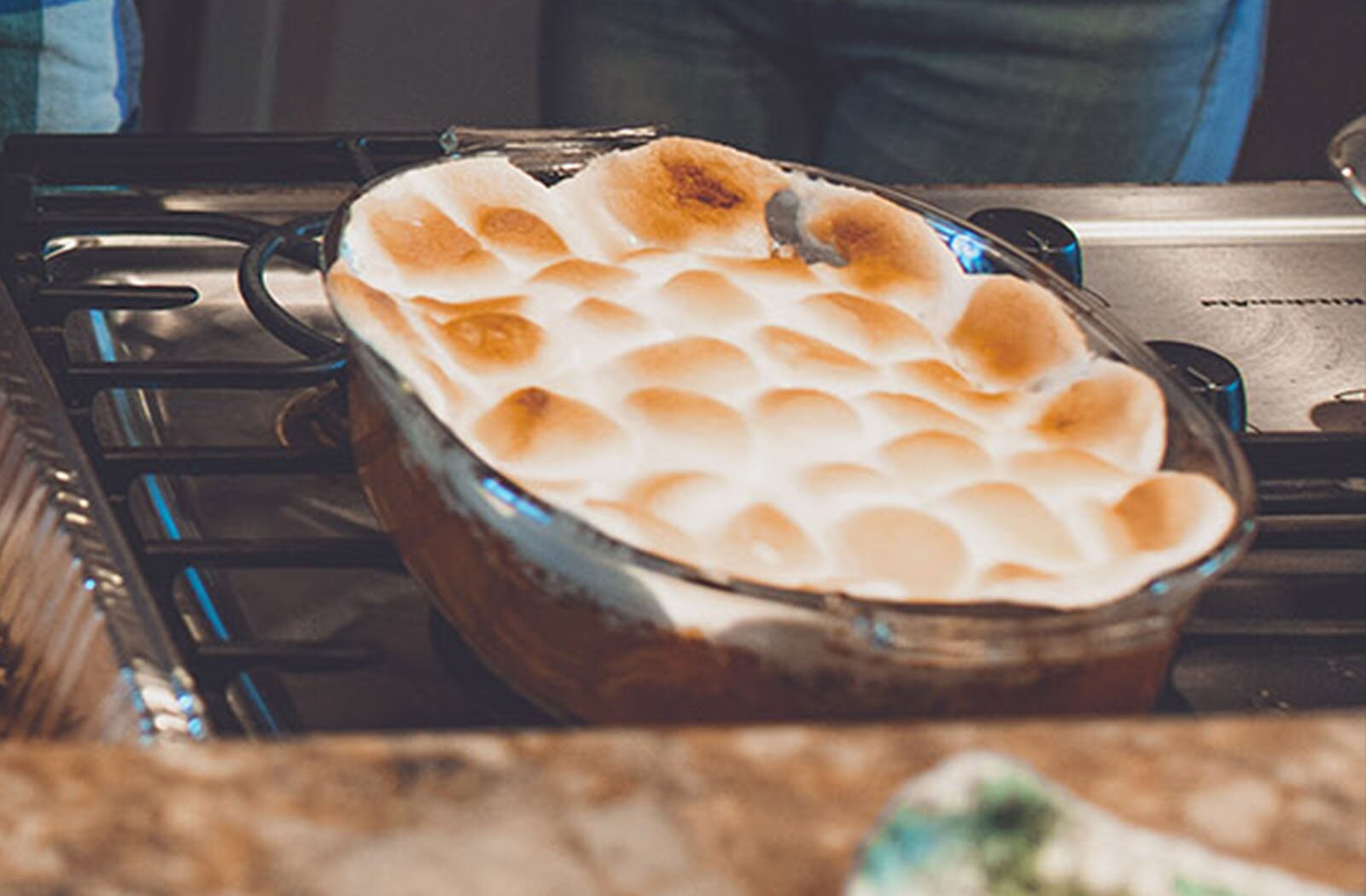 Zdjęcie przedstawia szklaną brytfankę wypełnioną zapiekanką, na wierzchu której widoczne są zapieczone pianki. Brytfanka stoi na ruszcie kuchni.