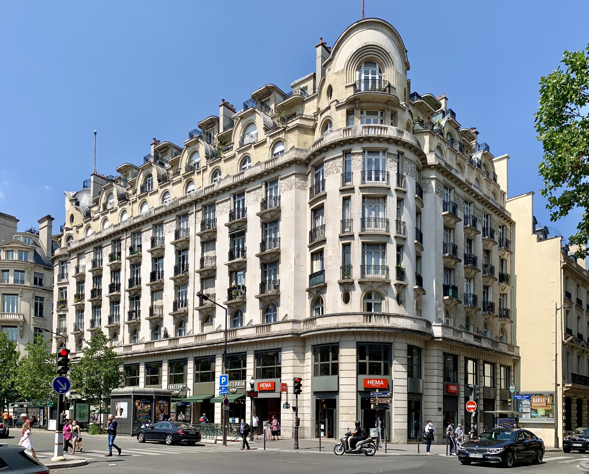 Fotografia dużych, monumentalnych kamienic o wyrafinowanym kształcie. W dolnej części wysokie okna przestrzeni usługowych, w górnej mieszkania z balkonami. Na pierwszym planie skrzyżowanie.