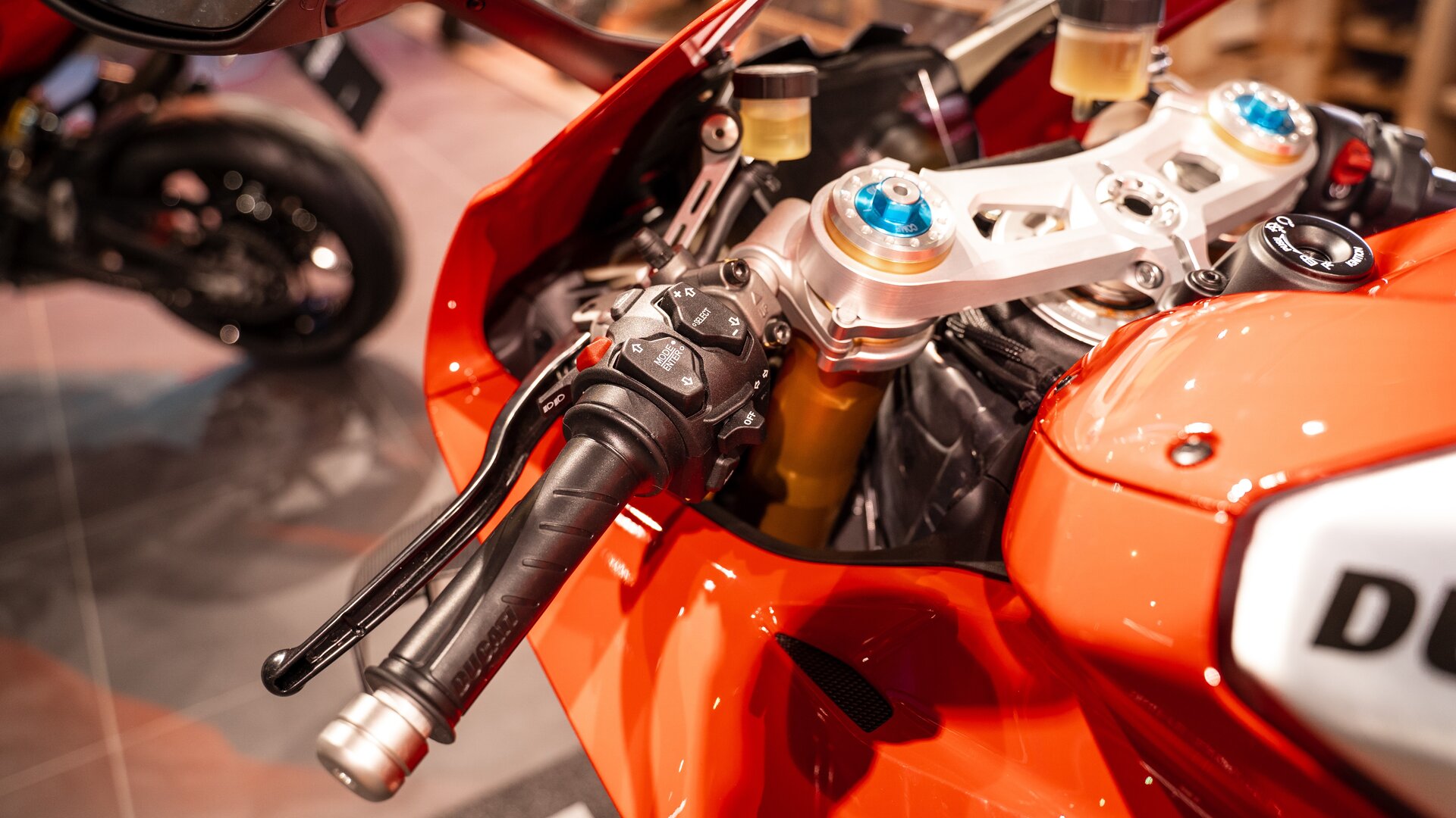 Na zdjęciu kierownica typu clip‑on. Jest ona elementem pomarańczowego motocyklu. Widać na niej hamulce, śrubki, a także dodatkowe przyciski służące do różnych funkcji.