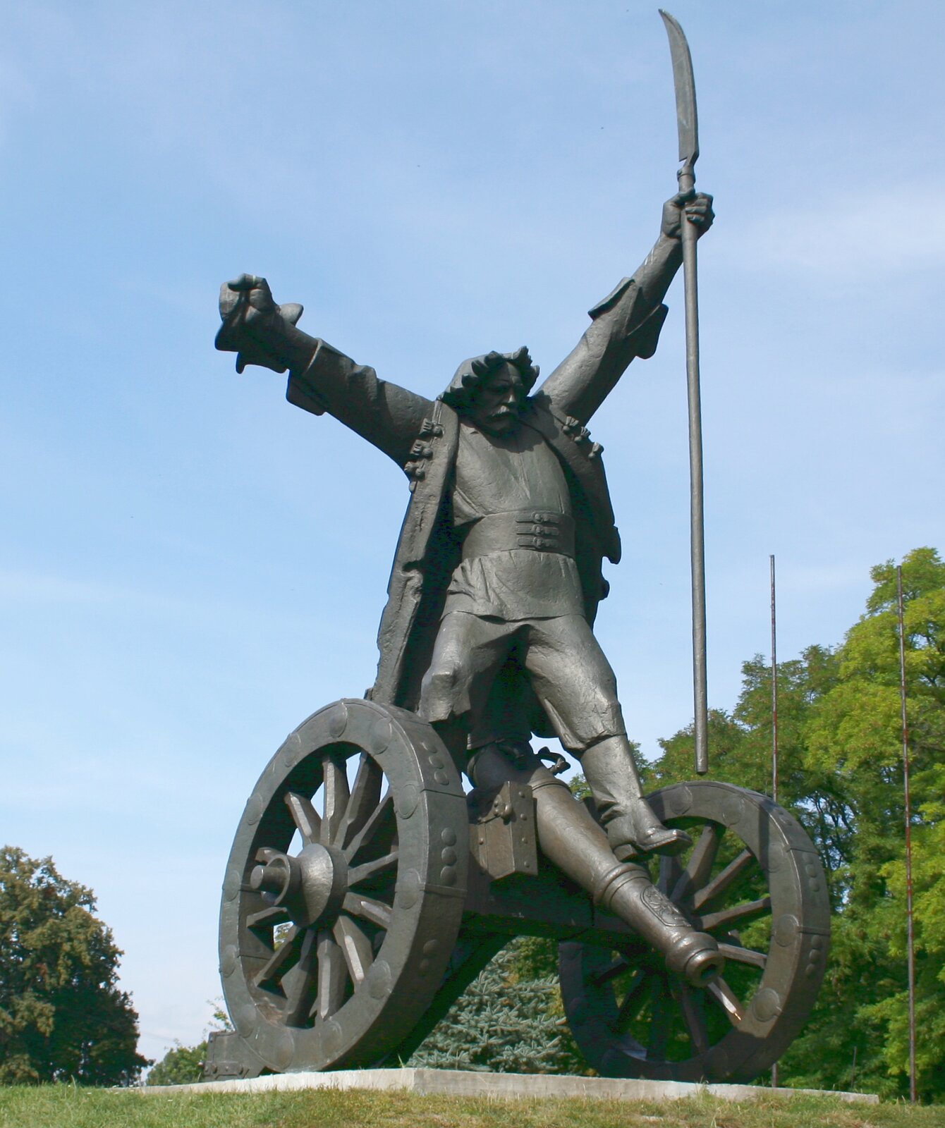 Fotografia przedstawia w ujęciu od dołu, na tle błękitnego nieba i szpaleru drzew, pomnik Bartosza Głowackiego w Janowiczkach. Bohater bitwy pod Racławicami został ukazany w długiej odrzuconej ku tyłowi chłopskiej sukmanie, z ozdobnym potrójnym zapięciem i rozciętymi podwiniętymi wysoko mankietami. Długa okrywająca biodra koszula jest w talii spięta szerokim pasem. Luźne chłopskie spodnie wpuszczone w buty z cholewami. 