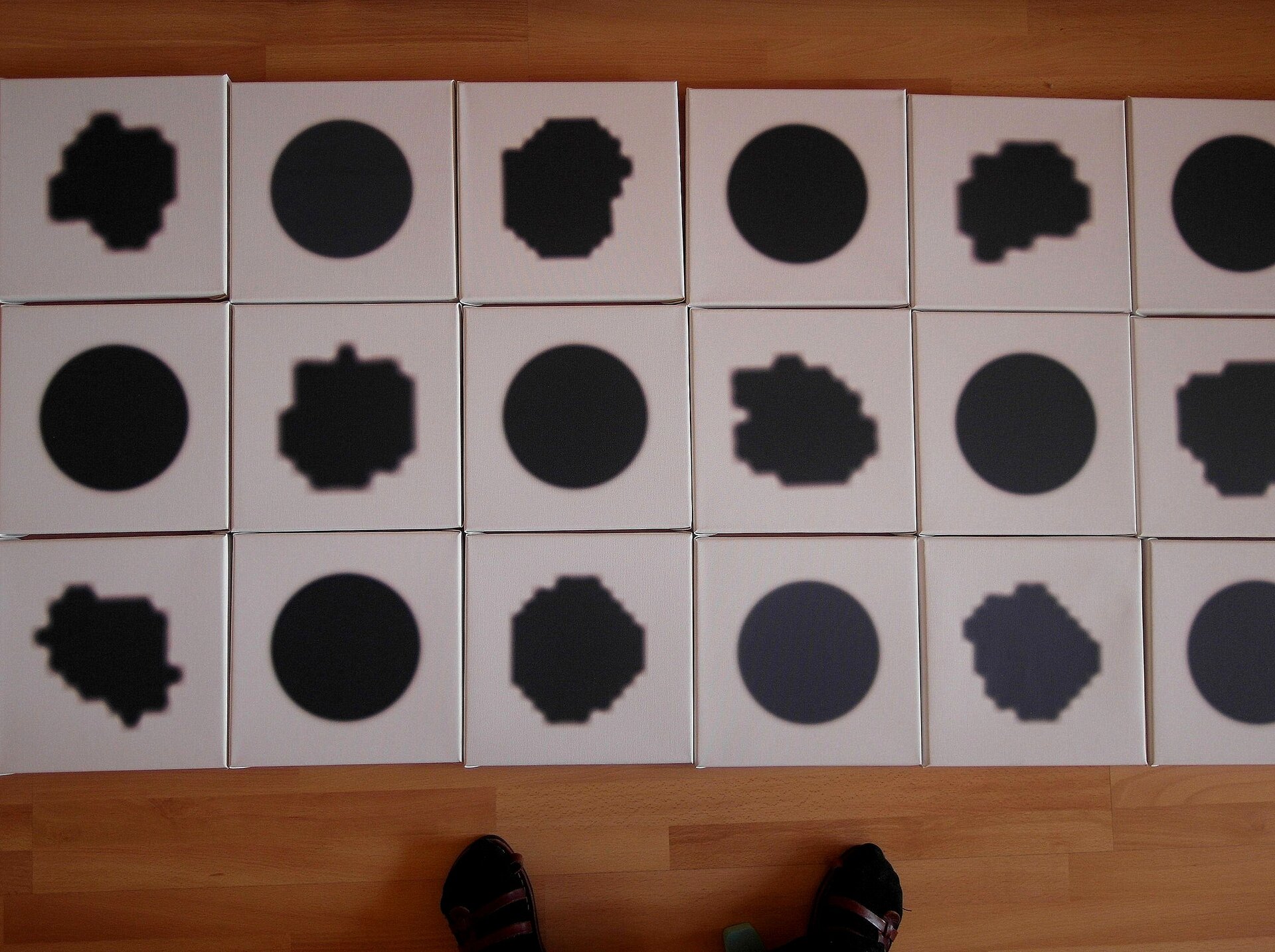 Ilustracja przedstawiająca rzut z góry na drewniana podłogę, na której rozłożone są obok siebie, w trzech rzędach jednakowej wielkości białe kwadraty. Na każdym z  nich jest jeden czarny punkt w dużym powiększeniu. Kwadraty z punktami są ułożone naprzemiennie: punkt wykonany komputerowo oraz punkt wykonany ręcznie. Punkty wykonane komputerowo są jednakowe, mają okrągły kształt. Punkty wykonane ręcznie mają nieregularny kształt, każdy jest inny.