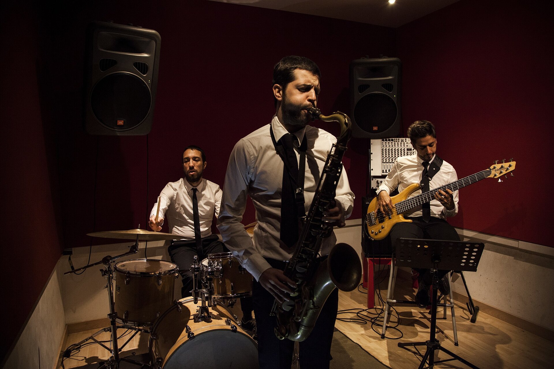 Zdjęcie przedstawia trzech mężczyzn grających na instrumentach w zamkniętym pomieszczeniu. Na pierwszym planie widoczny jest stojący mężczyzna w białej koszuli z czarnym krawatem, grający na saksofonie, który trzyma prawą ręką, zgiętą w łokciu, za lejkowato rozszerzającą się czarę głosową (roztrąb), zaś prawą ręką, zgiętą w łokciu, za klapy pionowego korpusu instrumentu. Po prawej stronie widoczny jest siedzący mężczyzna w białej koszuli z czarnym krawatem, grający na gitarze elektrycznej przewieszonej na czarnym pasku i korpusem spoczywającej na jego prawym biodrze. Jego prawa ręka, zgiętą w łokciu, spoczywa na korpusie gitary, zaś dłoń dotyka strun. Jego lewa dłoń dotyka gryfu gitary. Na lewo od stojącego mężczyzny widoczny jest siedzący mężczyzna w białej koszuli z czarnym krawatem, grający na perkusji. Jego prawa ręka jest wyprostowana, w dłoni trzyma pionową pałkę perkusyjną nad poziomo zorientowanym płaskim talerzem perkusyjnym. Poniżej talerza widoczne są dwa bębny, mniejszy o walcowatym kształcie w pozycji poziomej, większy – wysunięty do przodu, o walcowatym kształcie w pozycji pionowej. W tle, u góry, po prawej i po lewej stronie widoczne są dwa czarne prostopadłościany z zarysowanymi czarnymi kołami i prostokątami – obrazują one głośniki. Podłoga pomieszczenia jest w kolorze jasnobrązowym, zaś ściany pomieszczenia od podłogi są w kolorze jasnoszarym, natomiast od trzech czwartych są w kolorze brązowo‑czerwonym.