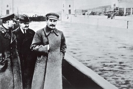 Kliknij, aby powiększyć Zdjęcie przedstawia sylwetki trzech mężczyzn. Wśród nich jest Józef Stalin. Ubrany jest w płaszcz, prawą rękę trzyma włożoną między zapięcia płaszcza, na wysokości klatki piersiowej. Pozostali mężczyźni są ubrani: pierwszy od lewej jest w wojskowy płaszcz, drugi w płaszcz. Obok przepływa woda, w tle widać zabudowania i most.