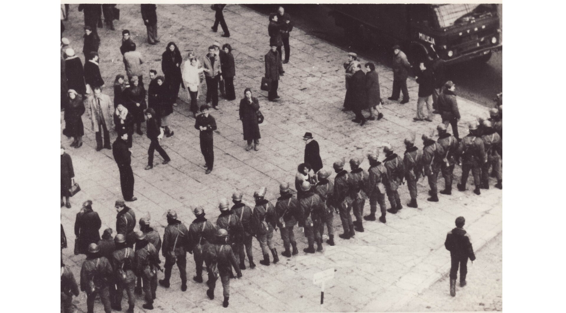 Czarno-biała fotografia przedstawiająca kordon funkcjonariuszy ZOMO w ubraniach bojowych i cywilów na chodniku.