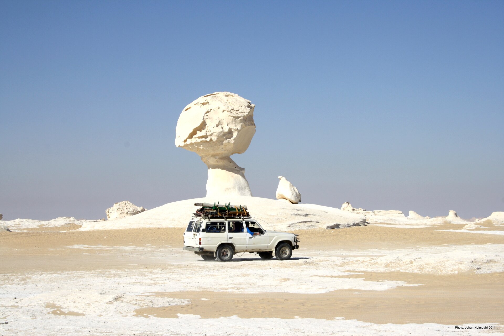 Na zdjęciu grzyb skalny na pustyni. Podstawa ma kształt walca, na jego czubku jest kula. Bardzo jasny kolor piasku. Na pierwszym planie płaski teren pustynny. Biały samochód terenowy, trzy razy mniejszy od grzyba.