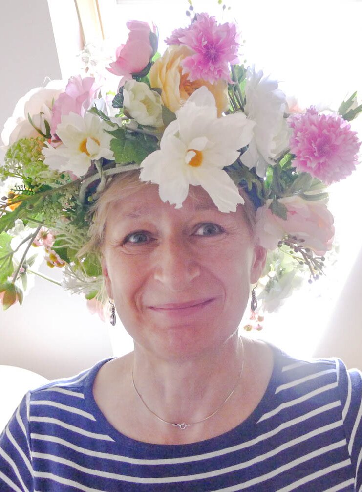 Fotografia przedstawia popiersie uśmiechniętej kobiety w średnim wieku, która ma na głowie wieniec z różnokolorowych kwiatów. Spod wianka widoczne są krótkie blond włosy. Ubrana jest w bluzkę w cienkie poziome paski. Nosi srebrne kolczyki i srebrny krótki łańcuszek. 