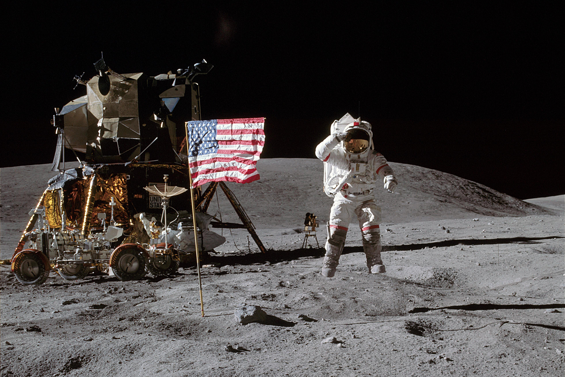 Fotografia prezentuje pierwszego człowieka na księżycu. Na pierwszym planie widoczny kosmonauta w białym kombinezonie stojący na powierzchni księżyca, po jego prawej stronie powiewa flaga amerykańska. Na lewo od kosmonauty widoczny pojazd, w tle czarne niebo.
