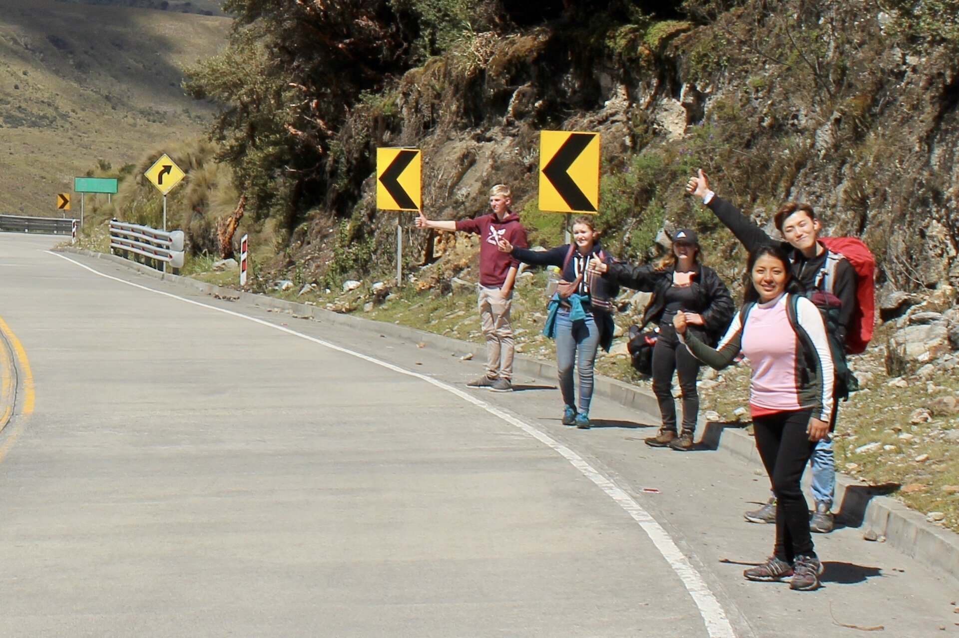 Zdjęcie przedstawia grupę pięciu młodych, uśmiechniętych osób: trzech dziewcząt i dwóch chłopców, ubranych w stroje i buty turystyczne. Młodzi ludzie stoją przy drodze wiodącej przez strome góry. Wszyscy mają plecaki. Wyciągają ręce z uniesionymi w górę kciukami w geście zatrzymywania autostopu.