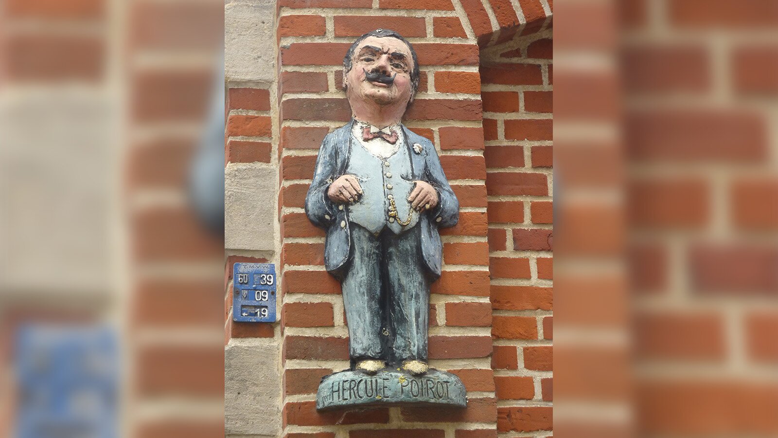 Zdjęcie przedstawia płaskorzeźbę na murze budynku z czerwonej cegły. Płaskorzeźba przedstawia mężczyznę z wąsem ubranego w kompletny garnitur. Pod jego stopami jest napis "Hercules Poirot". Transkrypcja znajduje się poniżej.