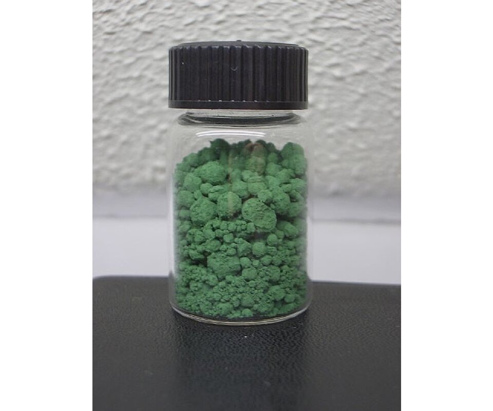 Zdjęcie przedstawia szklaną buteleczkę zamkniętą korkiem, w której przechowywane są granulki zielonego ciała stałego.