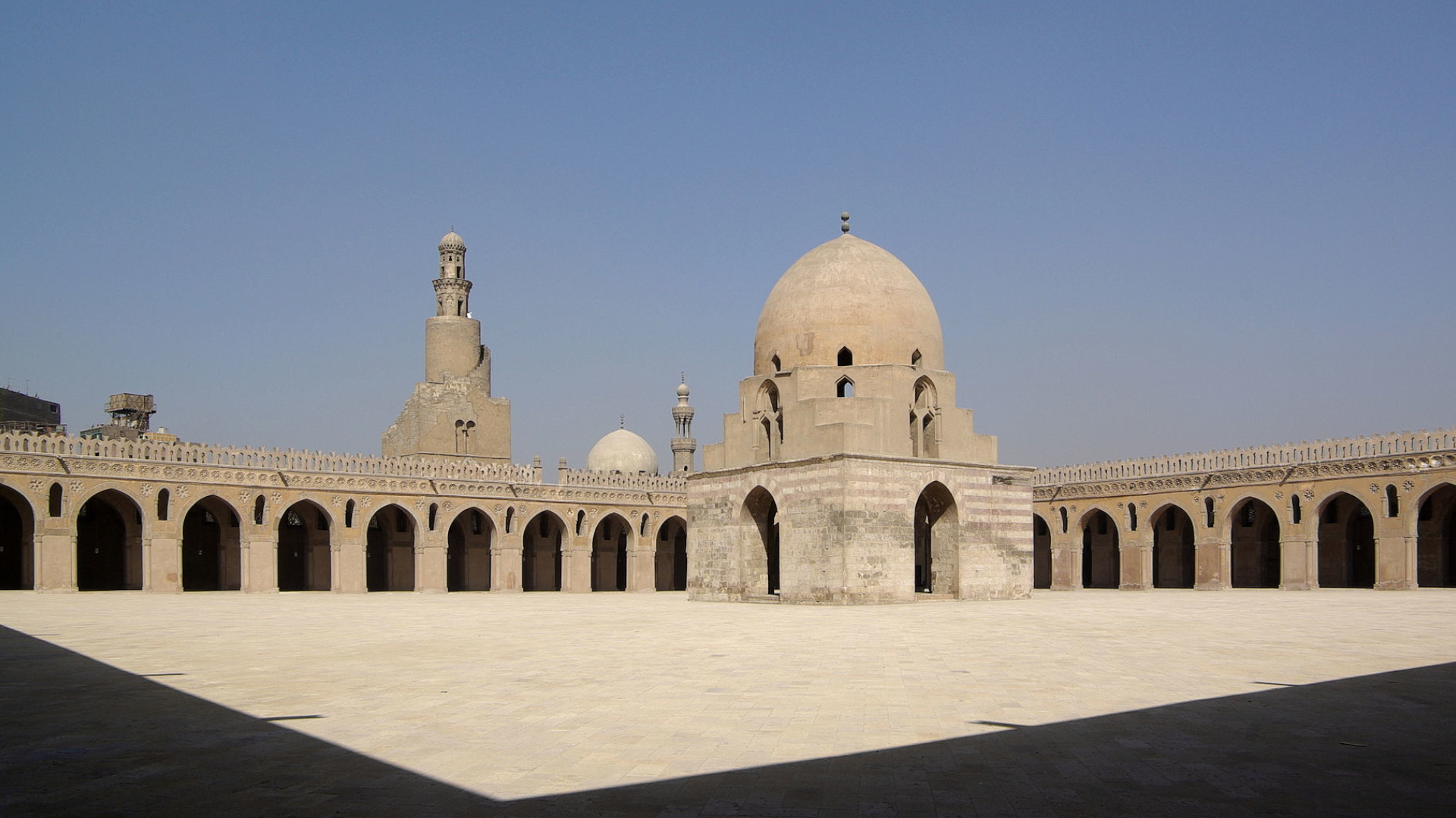 Na zdjęciu znajduje się dziedziniec meczetu, na którego środku znajduje się budowla z kopułą. Ściany meczetu składają się z łuków. W oddali znajduje się minaret oraz inne kopuły. 
