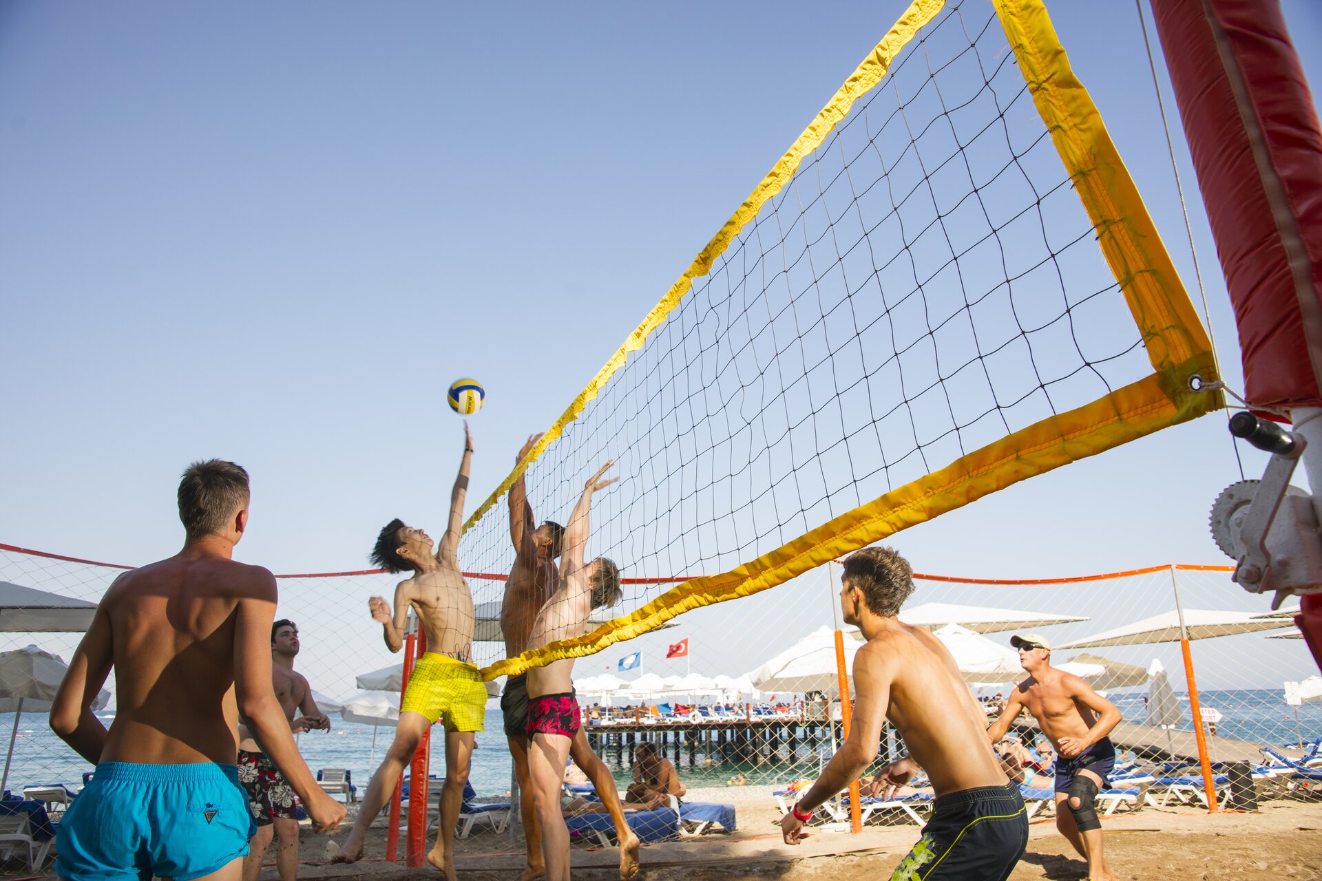 Zdjęcie przedstawia młodych mężczyzn grających w piłkę plażową. Widoczna jest rozpięta żółta siatka pomiędzy dwoma czerwonymi słupkami. Po lewej stronie siatki widoczne są trzy osoby, a po prawej cztery. Wszyscy mężczyźni są ubrani tylko w szorty w różnych kolorach. Chłopak stojący najbliżej siatki po lewej stronie podskakuje, ma lewą rękę wyciągniętą do góry i próbuje dotknąć piłkę znajdującą się tuż nad siatką. Dwóch chłopaków przy siatce po prawej stronie również podskakuje, ale ma wyciągnięte do góry obie ręce, które są ułożone do bloku. W tle widoczne są leżaki plażowe, na których siedzą ludzie, a dalej pomost z białymi parasolami oraz tafla niebieskiej wody.