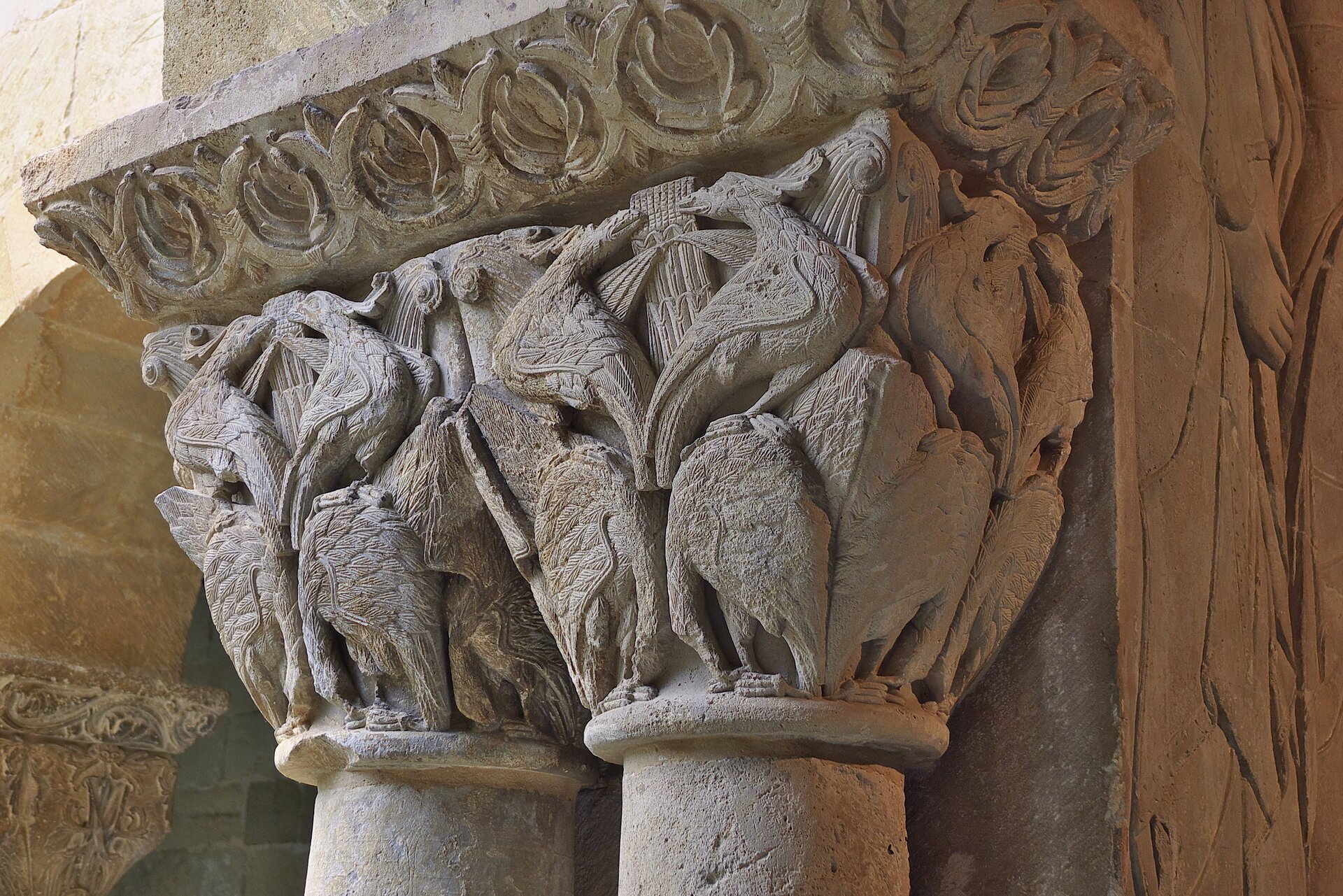 Fotografia przedstawia detal głowicy zoomorficznej, która ma kształt ściętego ostrosłupa. Głowica jest bogato zdobiona figurami ptactwa - część ptaków stoi (bez głów) a część siedzi na gałęzi.