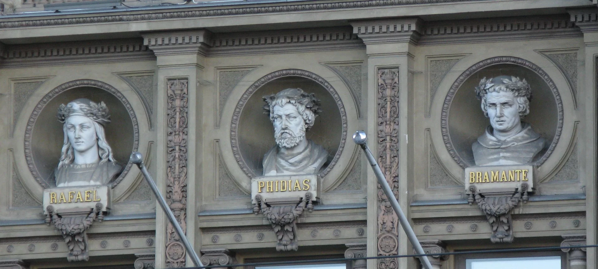 Zdjęcie przedstawia fragment ściany budynku. Obok siebie trzy wnęki w kształcie półkul. W każdej z nich umieszczono inne popiersie z podobizną mężczyzny. Popiersie po lewej strony, podpisane jest złotą czcionką: Rafael, kolejne: Phidias, a ostatnie: Bramante.