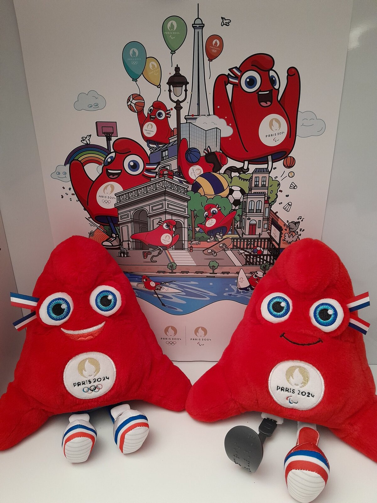 Na ilustracji znajdują się dwie czapki frygijskie - maskotki igrzysk olimpijskich i paraolimpijskich w 2024 roku. Siedzą oparte o ścianę, są pluszowe, mają kolor czerwony. Kształtem przypominają trójkąt – mają spiczasty, zaokrąglony czubek, który jest głową maskotki. Umieszczone są na niej duże, okrągłe, błękitne oczy, w ich zewnętrznych kącikach znajdują się dwie krótkie wstążki, posiadające trzy poziome pasy w kolorze niebieskim, białym i czerwonym – barwy flagi Francji oraz uśmiechnięte usta. Ich dwa rogi przy podstawie przypominają ręce. Maskotki posiadają cienkie nóżki oraz biało‑czerwono‑niebieskie buty. Większa maskotka nie ma jednej dolnej kończyny, zamiast tego posiada przytwierdzoną sportową protezę nogi. Obie mają na piersi białe koło z symbolem ognia olimpijskiego, napisem Paris 2024 i symbolem igrzysk olimpijskich przedstawiającym pięć złączonych pierścieni. Kręgi są splecione ze sobą w dwóch szeregach (od lewej do prawej): niebieski, czarny i czerwony na górze, żółty i zielony na dole. Za nimi widać kolorowy plakat promujący igrzyska z balonami, najważniejszymi zabytkami Paryża, rzeką Sekwaną i czerwonymi maskotkami.