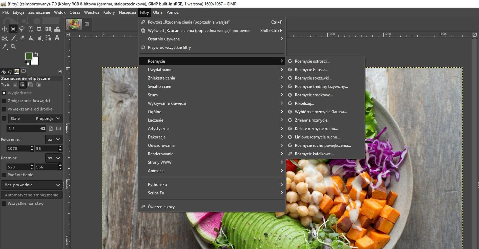 Ilustracja przedstawia okno programu. Po lewej stronie jest panel dotyczący Zaznaczenia eliptycznego. Z menu głównego wybrano zakładkę Filtry. Z listy wybrano opcję: Rozmycie. Przy Rozmyciu lista z różnymi rodzajami rozmycia, na przykład: Rozmycie ostrości, Rozmycie Gaussa, Rozmycie soczewki. W tle, w obszarze roboczym, jest zdjęcie pokrojonych warzyw. Znajdują się na talerzu.    