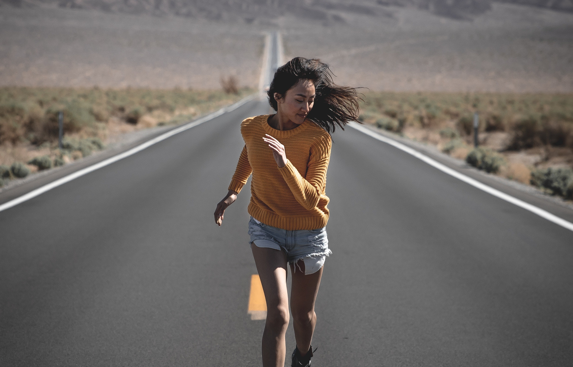 Zdjęcie przedstawia dziewczynę biegnącą środkiem asfaltowej szosy. Dziewczyna ma długie, ciemne i rozwiane włosy, jest ubrana w żółty sweter i dżinsowe spodenki. W tle za nią widoczna jest długa droga ciągnąca się przez pola porośnięte niewysoką i skąpą roślinnością.