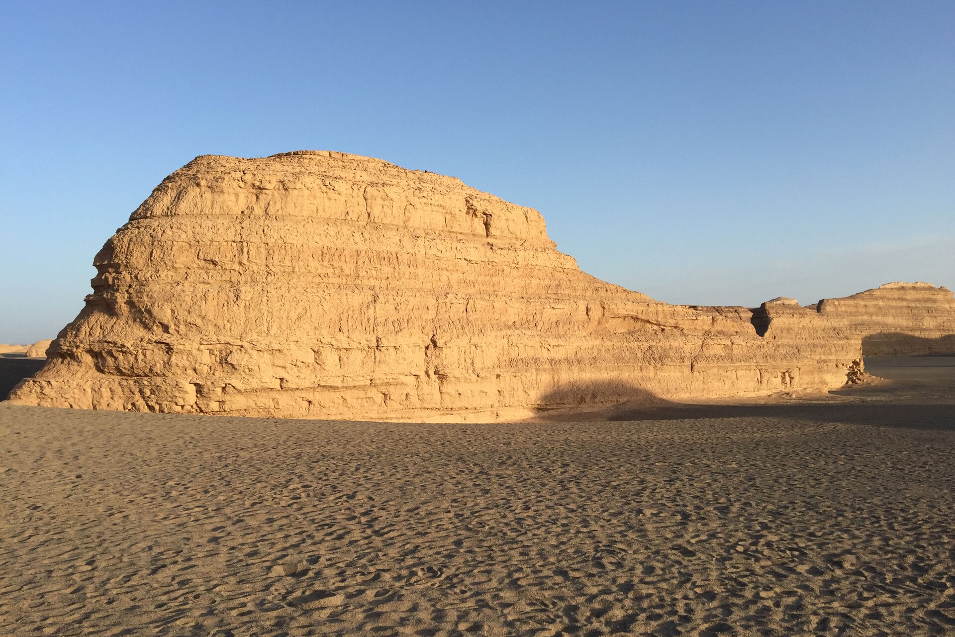 Na zdjęciu Jardang. Szczyty spłaszczone, ściany strome. Na pierwszym planie płaski, piaszczysty teren. Powyżej bezchmurne niebo. Po lewej stronie, na ścianie Jardangu widoczne poziome żłobienia.