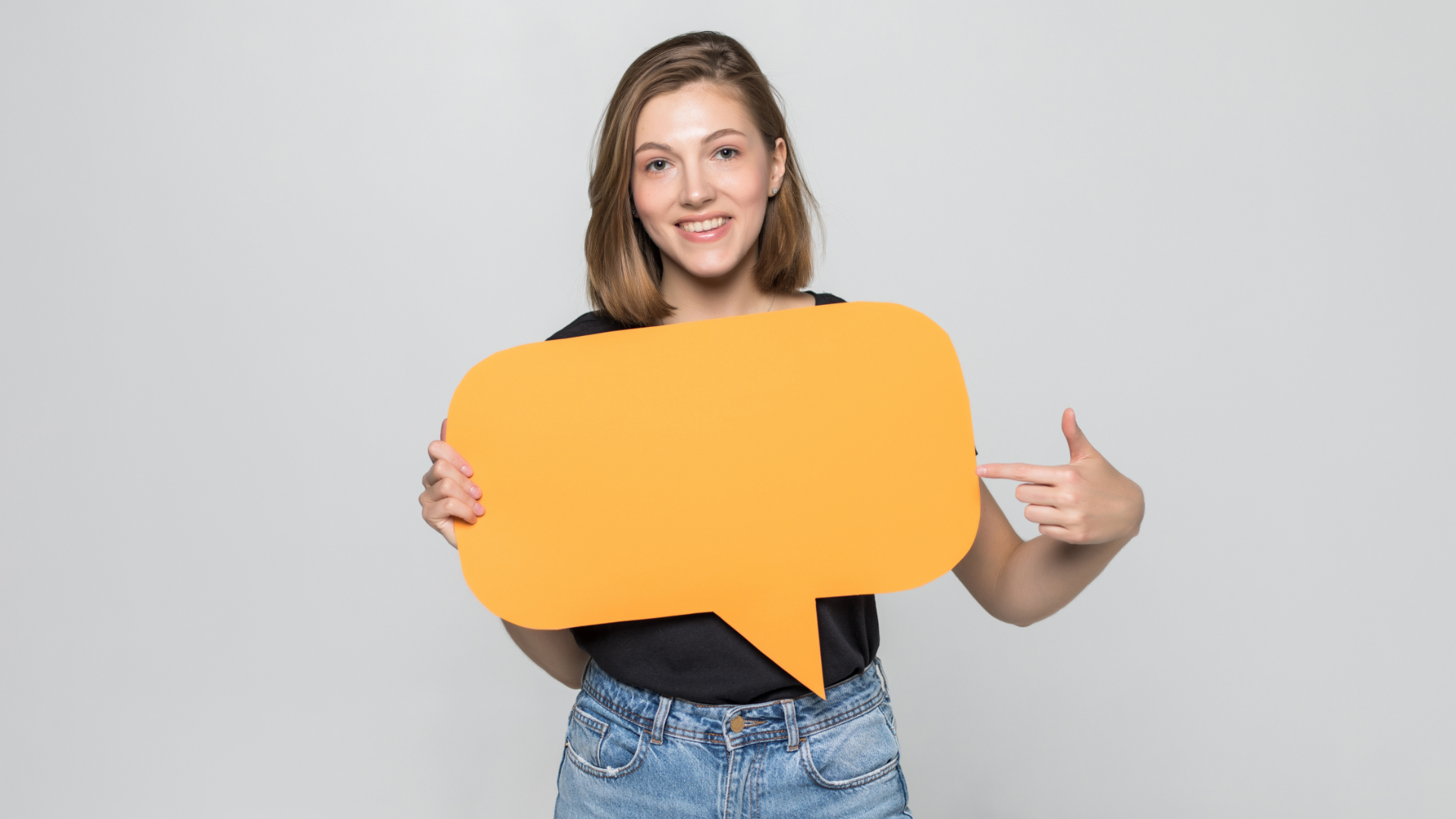 Zdjęcie przedstawia młodą, uśmiechniętą. dziewczynę z prostymi włosami do ramion. Dziewczyna w dłoniach trzyma dużą, papierową ikonę czatu/dymku rozmowy. Dymek jest pusty, nie ma na nim tekstu.