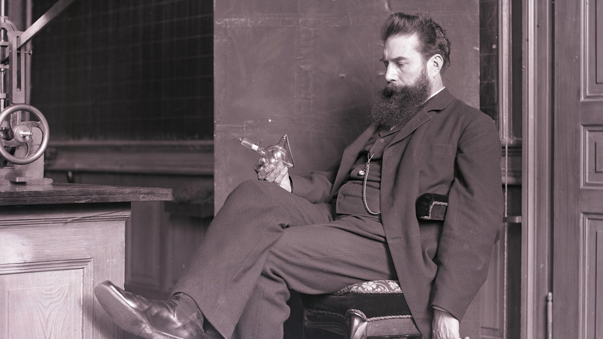 Rys. 4. Zdjęcie przedstawia brodatego mężczyznę siedzącego w swobodnej pozie przy stole z aparaturą. Mężczyzna spogląda na szklaną bańkę (lampę rentgenowską), którą trzyma w prawej dłoni opartej o kolano. 