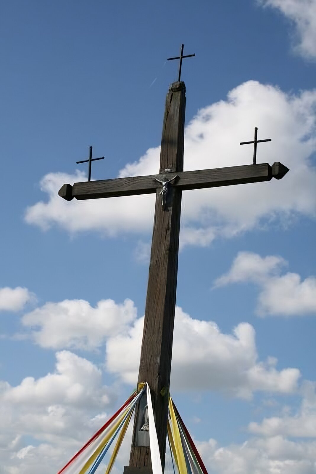 Zdjęcie przedstawia drewniany krzyż, u którego na czubkach ramion umieszczone zostały kolejne drewniane krzyże. U dołu krzyża przywiązane zostały szarfy w kolorze: białym, czerwonym, żółtym, niebieskim. W tle widoczne jest błękitne niebo, częściowo przysłonięte białymi chmurami. Zdjęcie zostało wykonane w świetle dziennym.