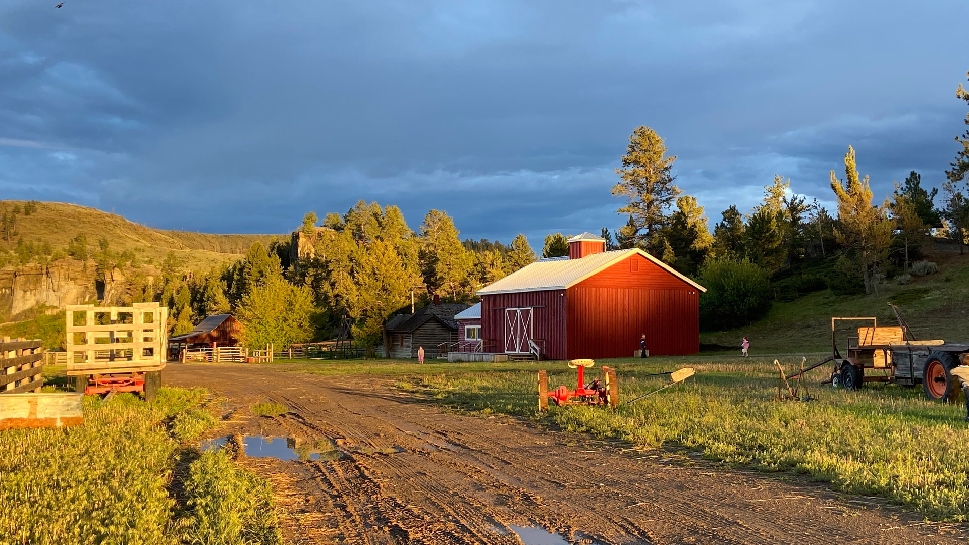 Zdjęcie przedstawia wiejskie gospodarstwo w świetle zachodzącego słońca. Po prawej stronie widoczne są budynki gospodarstwa i kilka maszyn rolniczych. Do gospodarstwa prowadzi zabłocona droga z kałużami. Po lewej stronie stoi przyczepa.