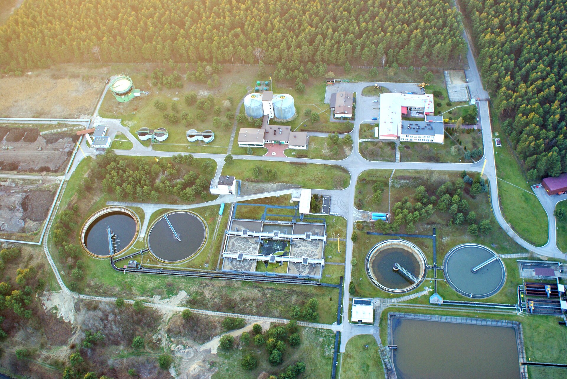Na zdjęciu oczyszczalnia ścieków. Zdjęcie z góry. Duże okrągłe zbiorniki. Wewnątrz mieszadła zbierające nieczystości. Budynki techniczne. Drogi. Na drugim planie las.