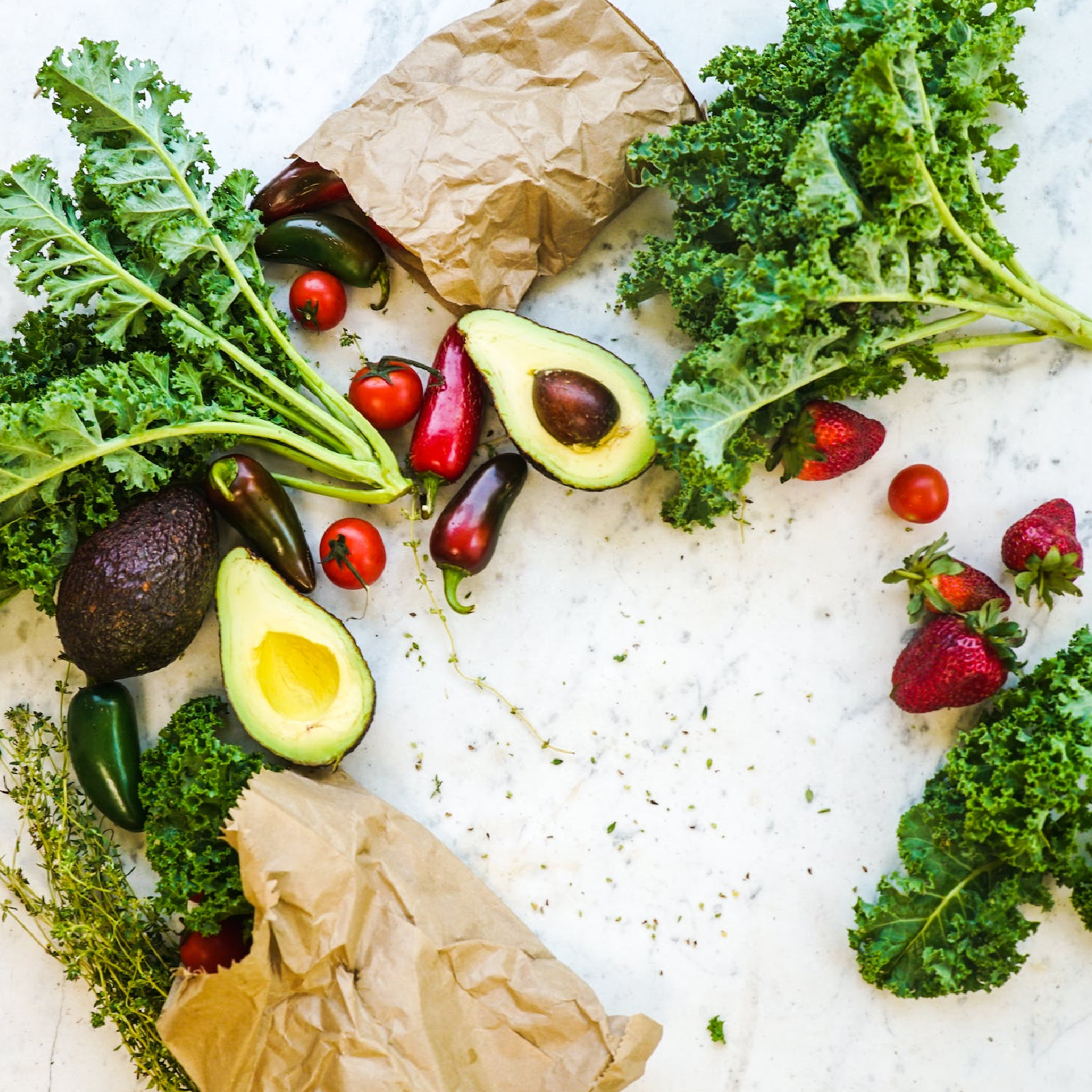Zdjęcie przedstawia owoce i warzywa, rozsypane na stole. Widoczne jest awokado przecięte na pół, truskawki, papryczki, pomidory, i inne zielone warzywa.