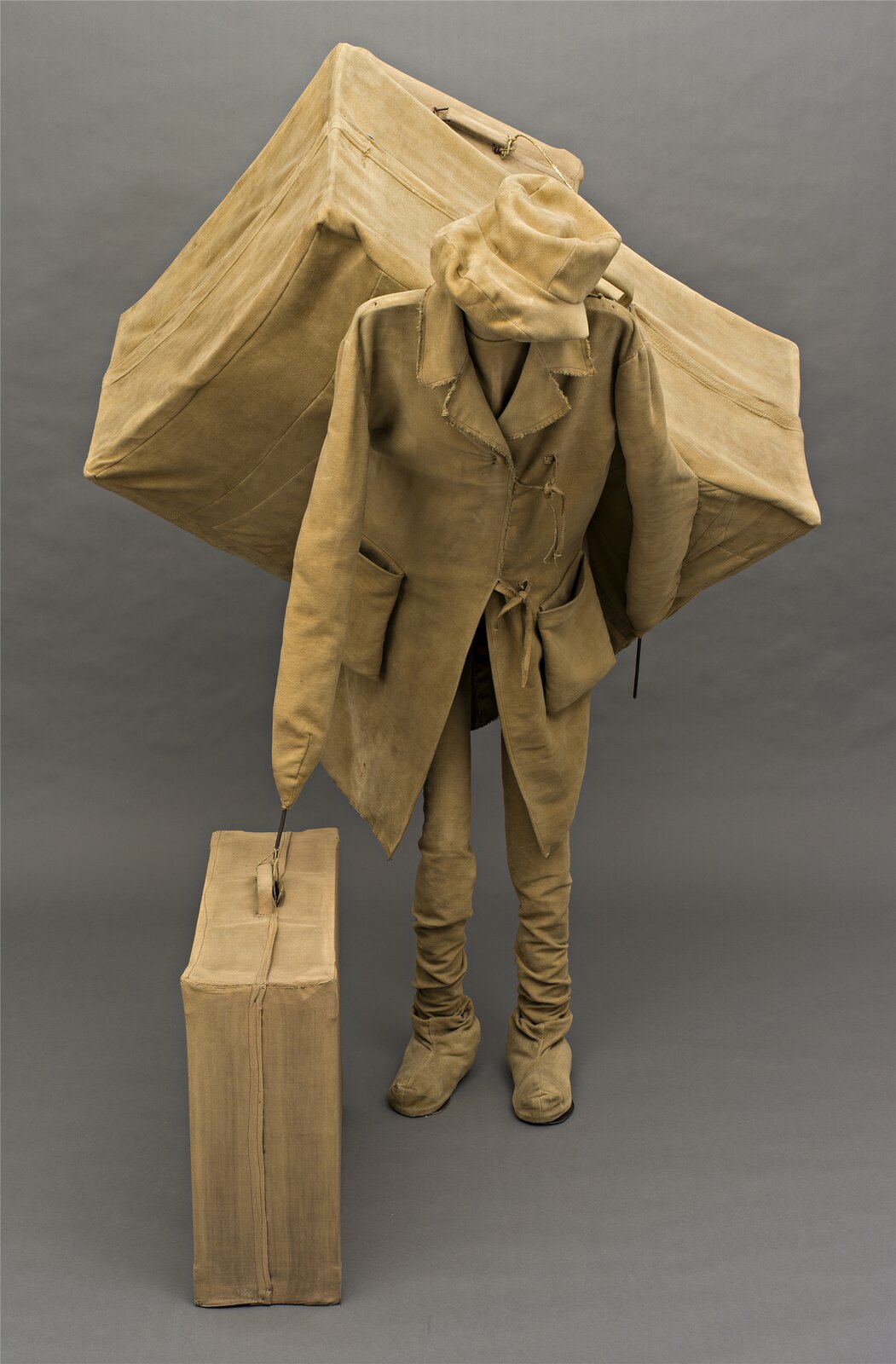 Rzeźba przedstawia sylwetkę mężczyzny ubranego w buty o wysokich cholewkach, luźny, długi płaszcz oraz kapelusz. Materiał butów jest pomarszczony, a kapelusz oklapnięty. Pod prawym rękawem płaszcza widoczna jest duża walizka, a na plecach mężczyzny – wielka skrzynia z przymocowaną do niej mniejszą walizką. Sylwetka postaci jest pochylona, a głowa skierowana w dół. Cała rzeźba, przedstawiona na szarym tle, utrzymana jest w tym samym, wyblakłym odcieniu koloru żółto‑brązowego.