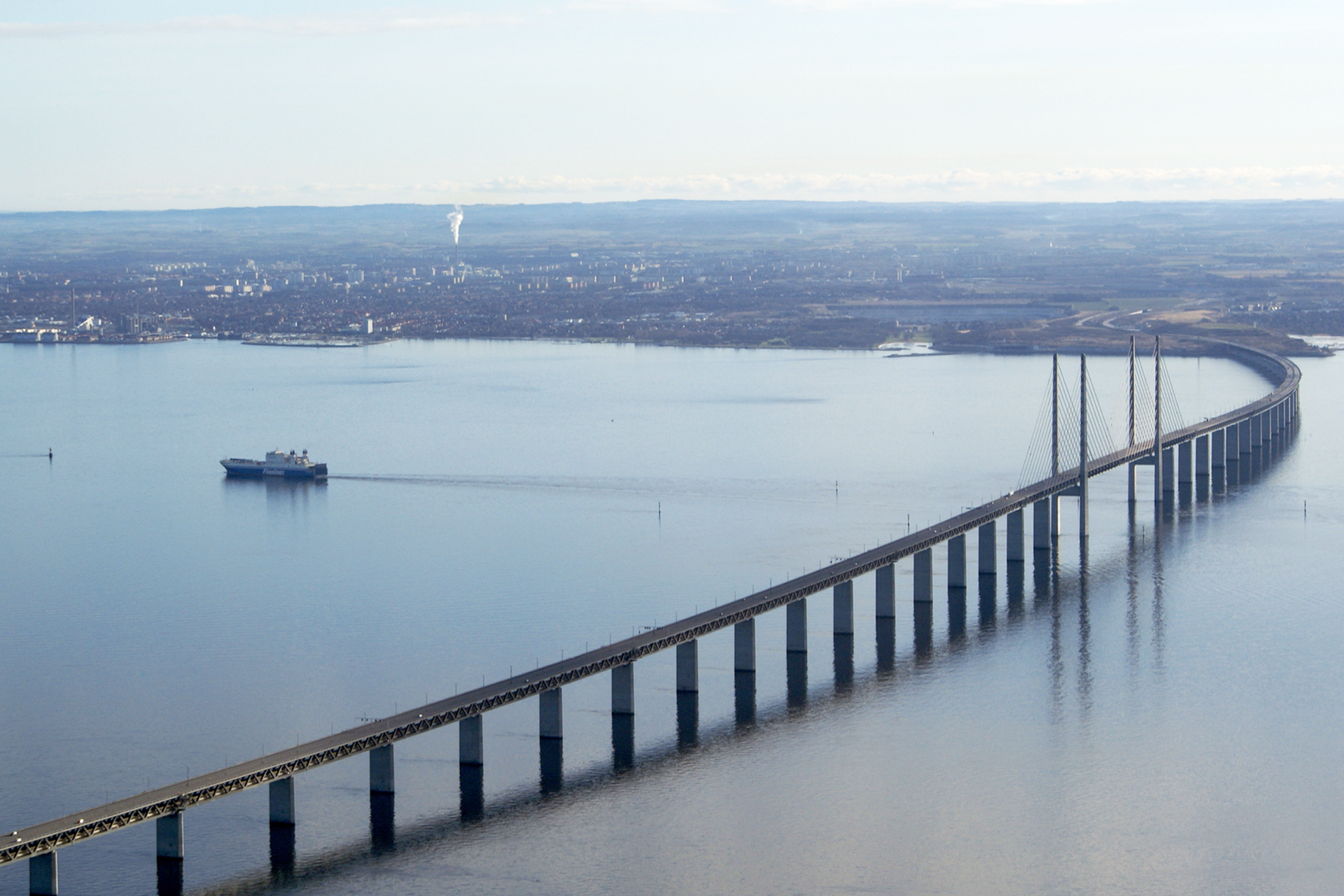 Fotografia przedstawia długi, przęsłowy most łączącego dwa odległe brzegi. Most znajduje się nad cieśniną Sund. Jest wysoki, widać, że przepłynął pod nim duży statek. W tle widoczny ląd, na którym znajduje się miasto.
