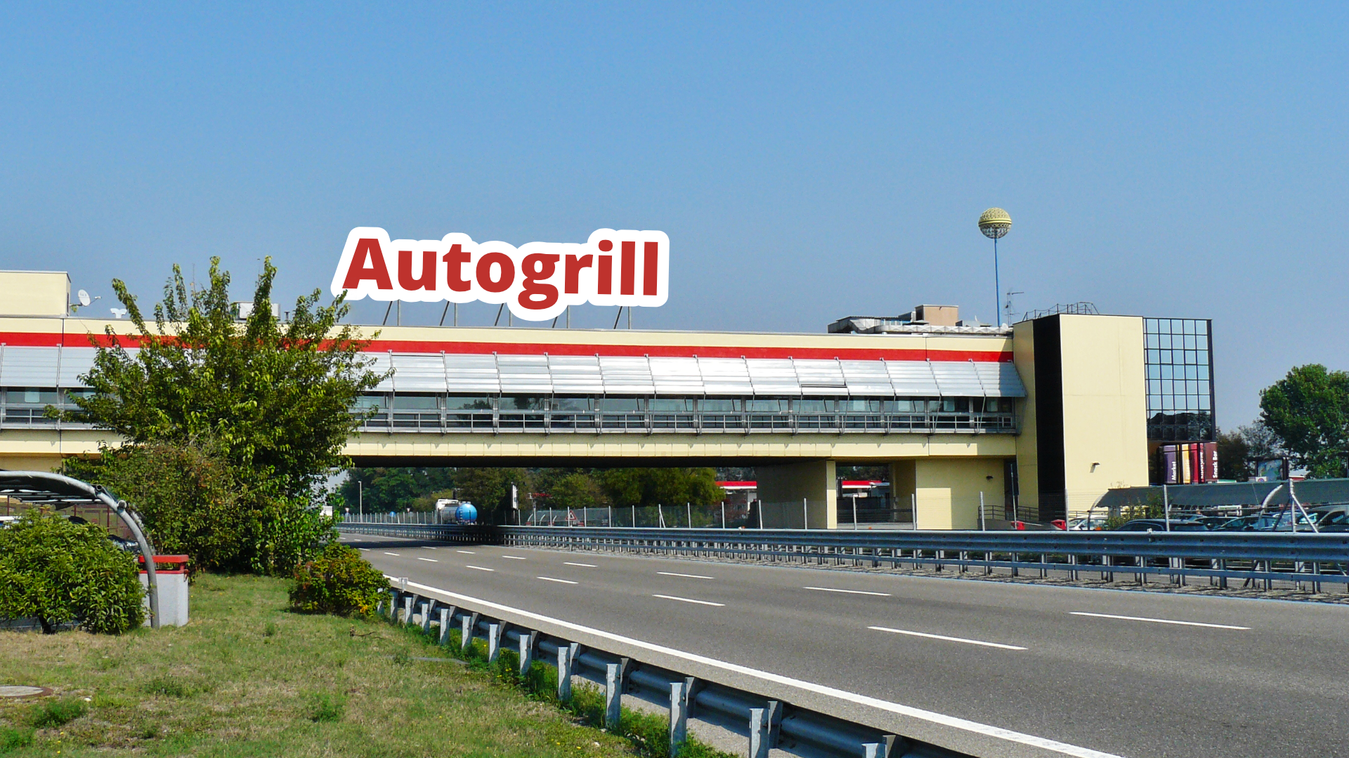Zdjęcie przedstawia fragment autostrady z kilkoma pasami ruchu. Nad autostradą znajduje się budynek z przeszklonymi szybami. Nad budynkiem widoczny jest napis: "Autogrill".