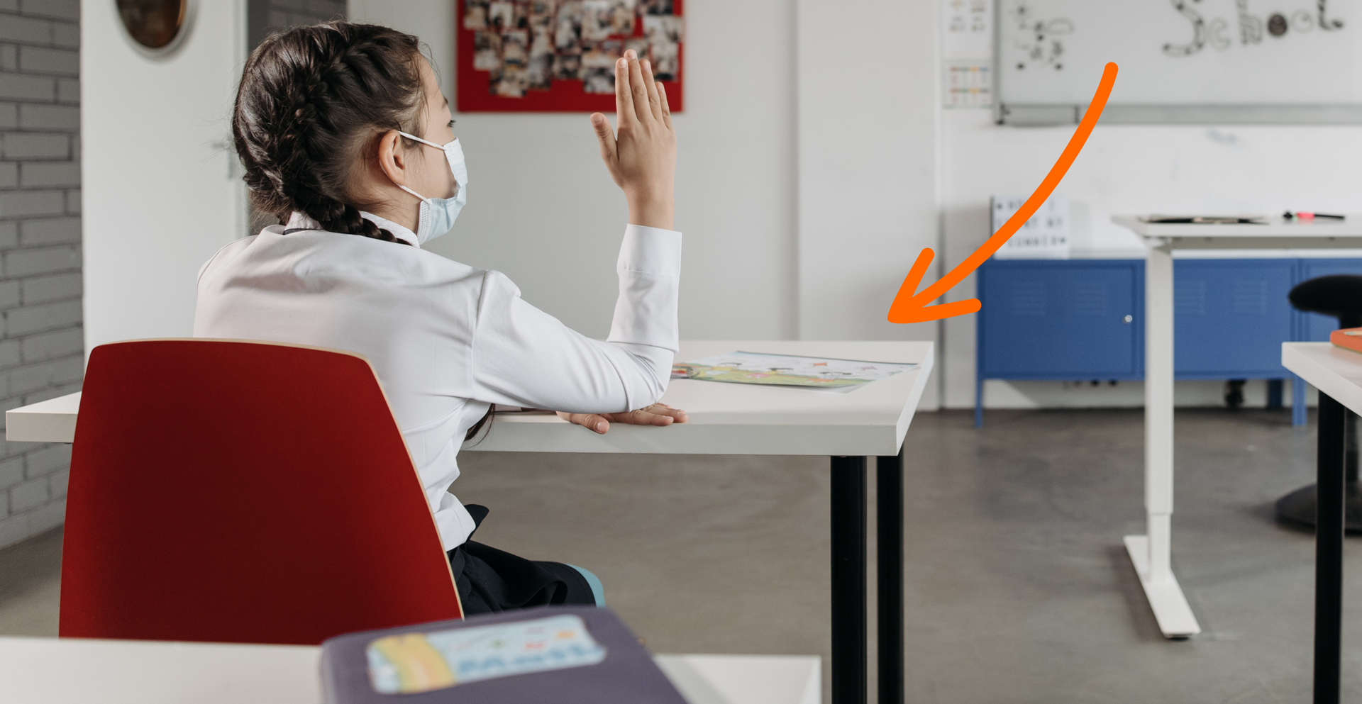 Zdjęcie przedstawia uczennicę siedzącą w ławce szkolnej. Dziewczynka siedzi tyłem do kadru, podnosi prawą rękę, nosi ochronną maseczkę. Na zdjęciu znajduje się strzałka skierowana na ławkę.