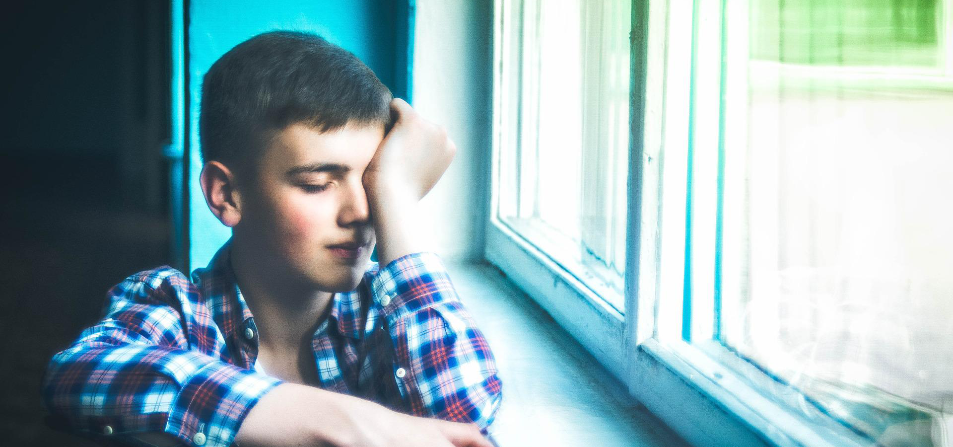 Zdjęcie przedstawia chłopca ubranego w koszulę w kratkę. Znajduje się przy oknie i opiera ręką głowę.