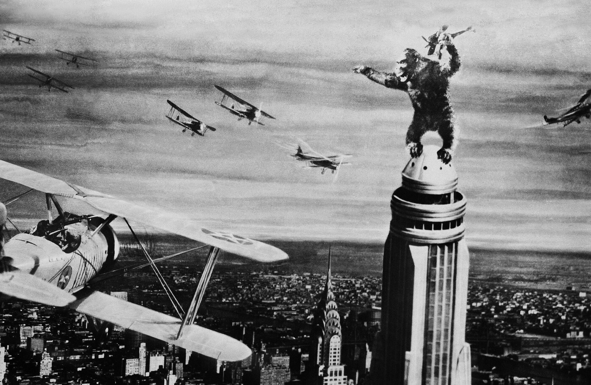 Czarno‑biała fotografia przedstawia kadr z filmu King‑Kong. Na pierwszym planie drapacz chmur Empire State Building, na szczycie którego stoi ogromny goryl trzymający w ręku samolot. W jego kierunku zbliżają się inne samoloty. W tle zabudowania miasta.