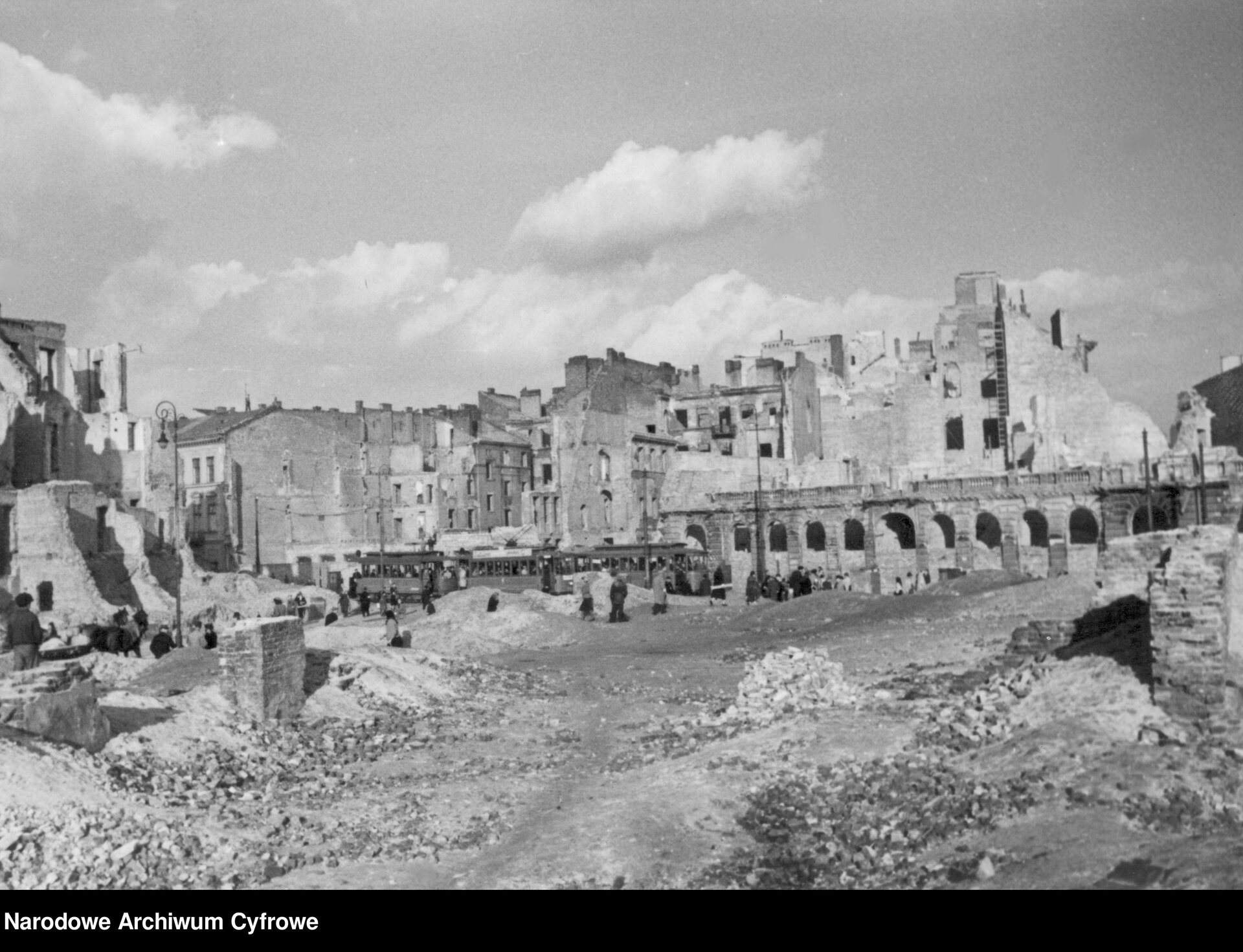 Zdjęcie przedstawia Warszawę po wojnie. Na zdjęciu z Narodowego Archiwum Cyfrowego są całkowicie lub częściowo zburzone budynki. Brak ulic, widać tylko gruzy oraz gdzieniegdzie stojących ludzi i tramwaj pełen ludzi.   