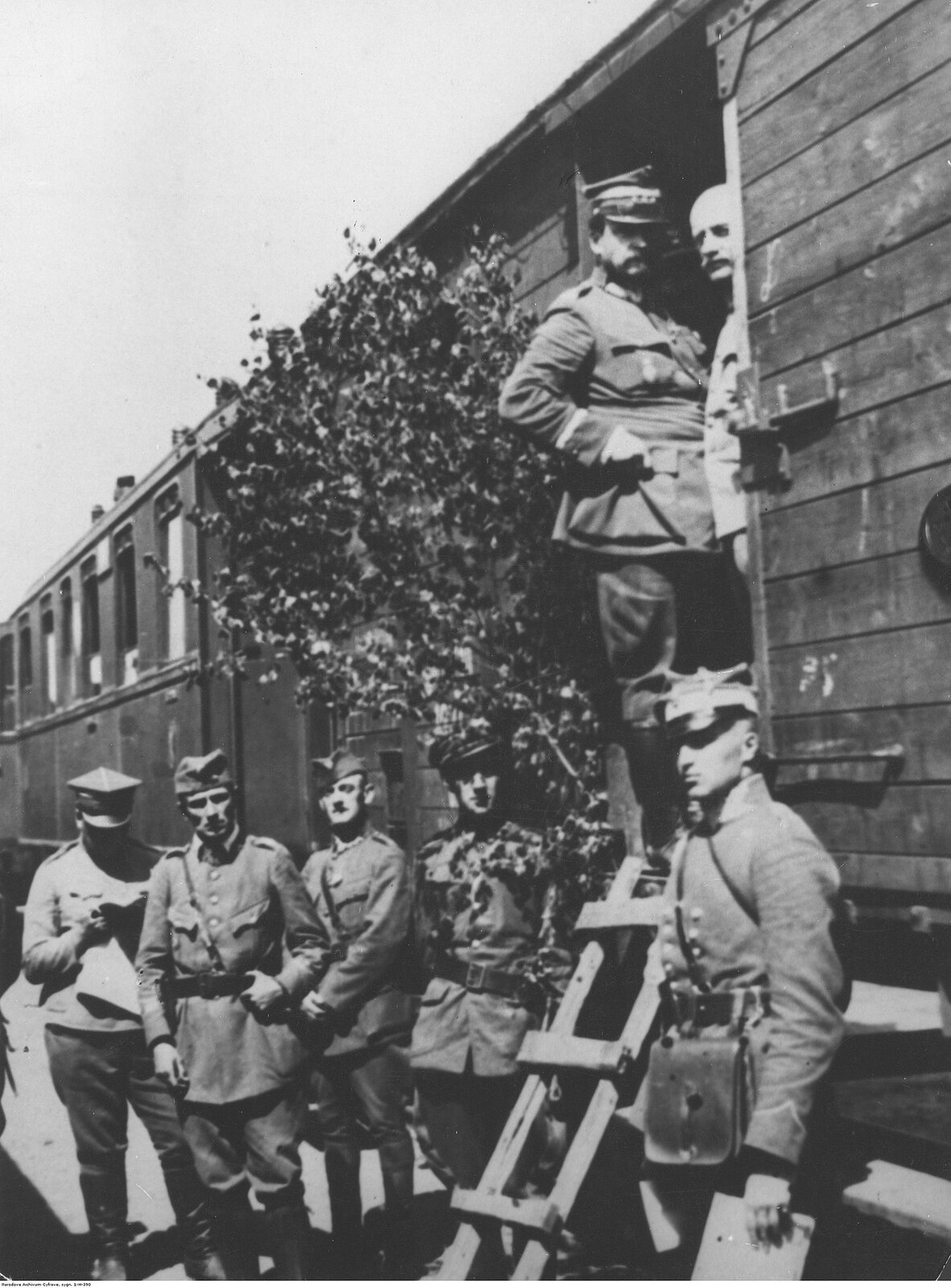 Zdjęcie przedstawia wysokiego rangą żołnierza polskiego stojącego na progu otwartego wagonu towarowego. NA głowie ma czapkę rogatywkę z kwadratowym denkiem. Długie, sumiaste wąsy. W progu wagonu stoi także inny mężczyzna, a na peronie przy wejściu do wagonu grupa szeregowych żołnierzy w polskich mundurach.