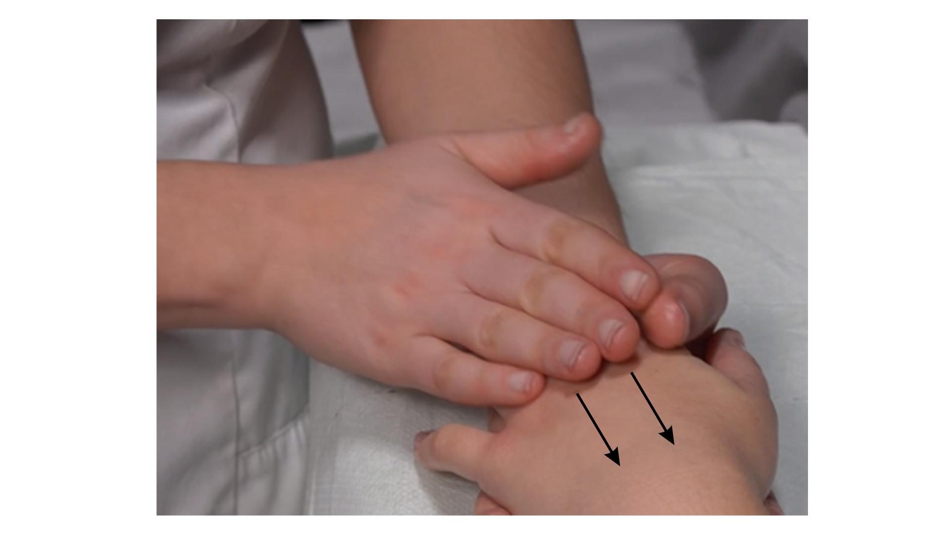 Zdjęcie przedstawia lewą rękę pacjenta leżącą na stole do masażu. Masażysta wykonuje ruch liniowy opuszkami palców na grzbiecie ręki pacjenta od strony palców w stronę nadgarstka. 