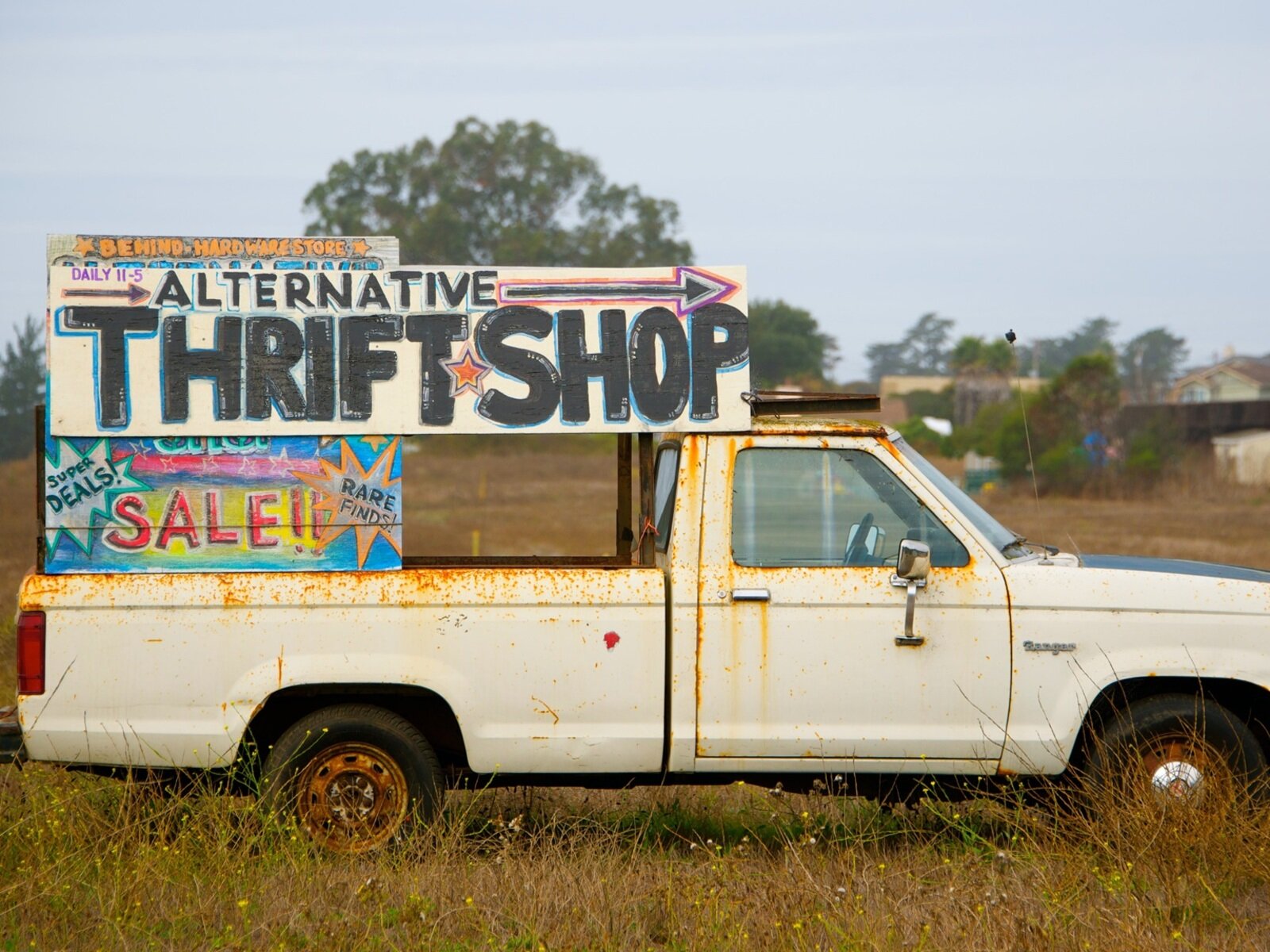 Na zdjęciu widoczny jest stary, ociekający rdzą samochód w typie pickup w kolorze białym. Na nim umieszczono tablicę z napisem w języku angielskim ALTERNATIVE THRIFT SHOP. Oraz tabliczkę z napisem SALE. Samochód stoi na łące w szczerym polu. 