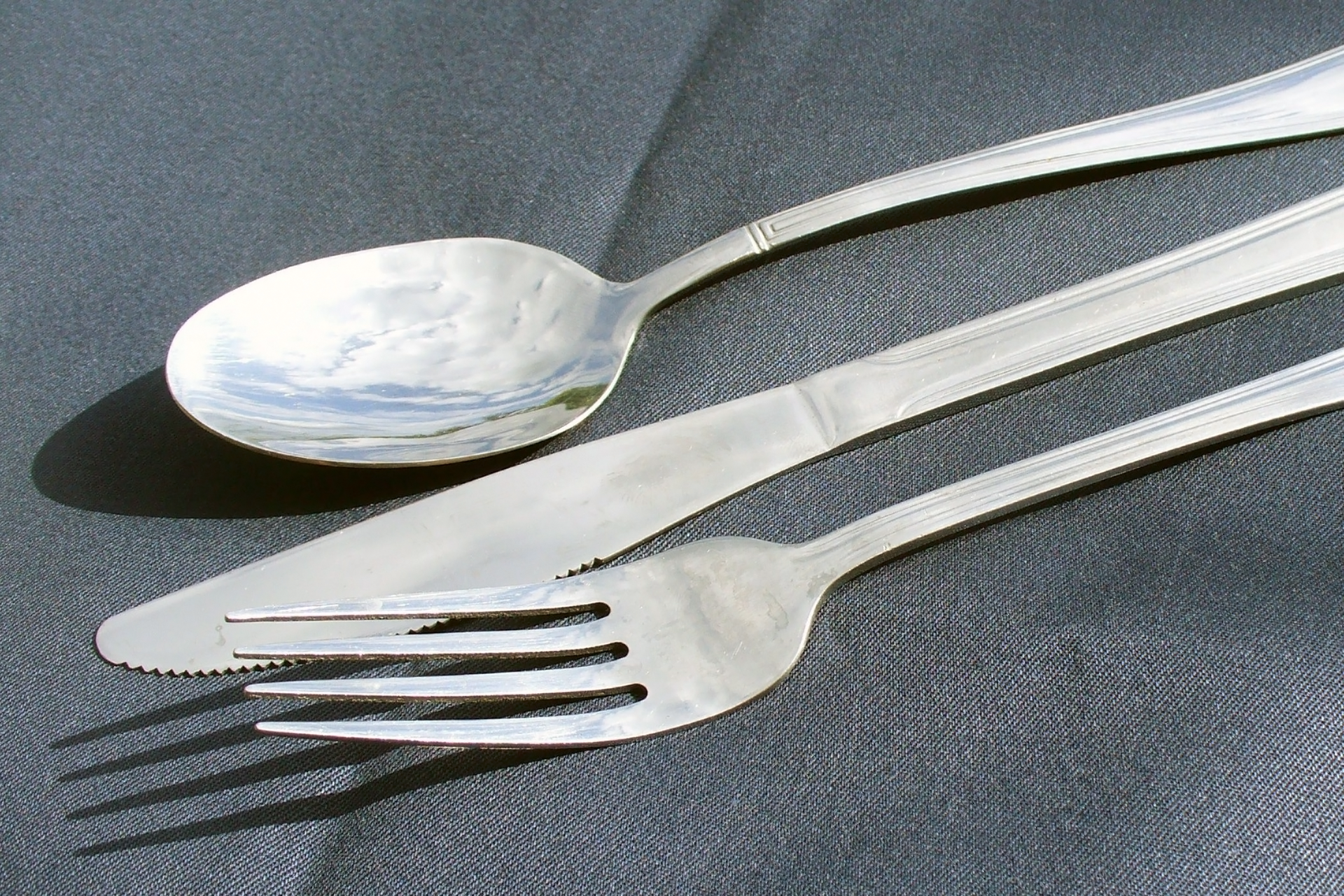 Fotografia przedstawia metalowe sztućce: łyżkę, nóż i widelec na szarym materiale. Na sztućcach widoczny jest charakterystyczny dla metalu połysk.