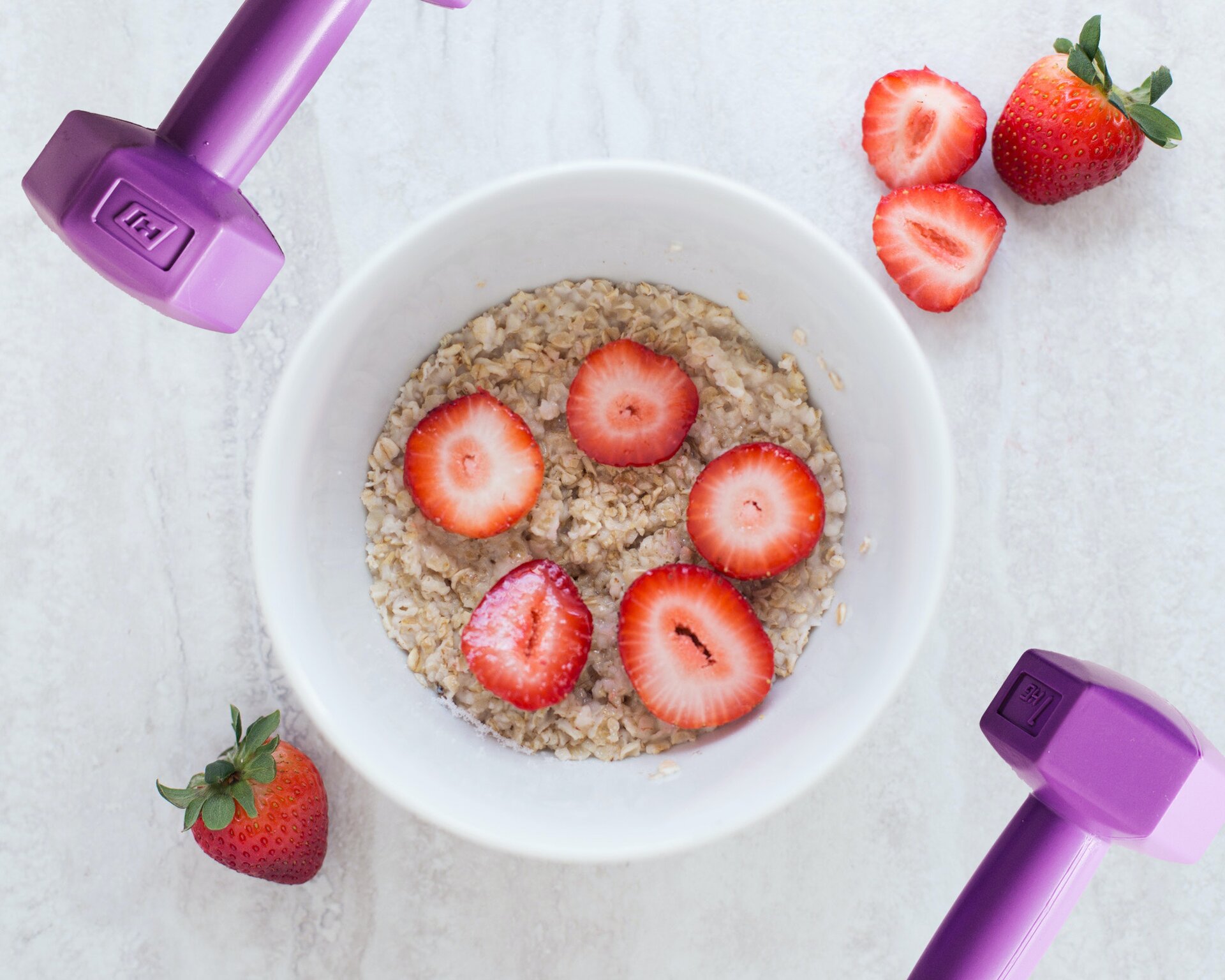 Zdjęcie przedstawia białą miseczkę, w ujęciu z góry, stojącą na szarym blacie. W miseczce widoczne są płatki owsiane oraz połówki truskawek ułożone w kółko. Po prawej i lewej stronie miseczki widoczne są fioletowe, średniej wagi hantle oraz całe i przekrojone truskawki.