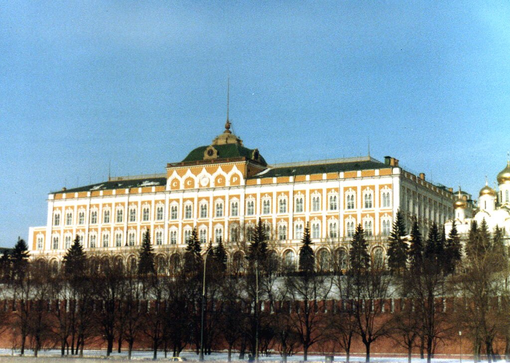 Na zdjęciu znajduje się Wielki Pałac Kremlowski. Jest to ogromny budynek o złoto białych ścianach i zielonym dachu. Posiada on liczne okna. W jego głównej części widoczne są złote zdobienia i iglica. Znajdują się w nich symbole komunistyczne. Przed pałacem widoczny jest park, ceglane mury obronne i rzeka. 