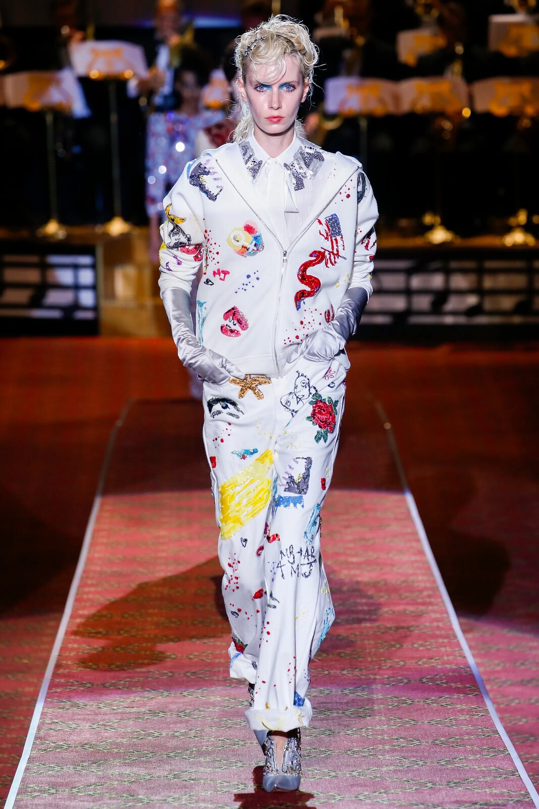 Fotografia przedstawia modelkę na wybiegu. Ma ona  na sobie biały, luźny kombinezon we wzory przedstawiające między innymi czerwonego węża, żółtą rozgwiazdę oraz szare napisy. Buty oraz rękawiczki kobiety są srebrne. Jej makijaż oczu utrzymany jest w odcieniach niebieskiego oraz różu, a jej blond włosy są spięte. 