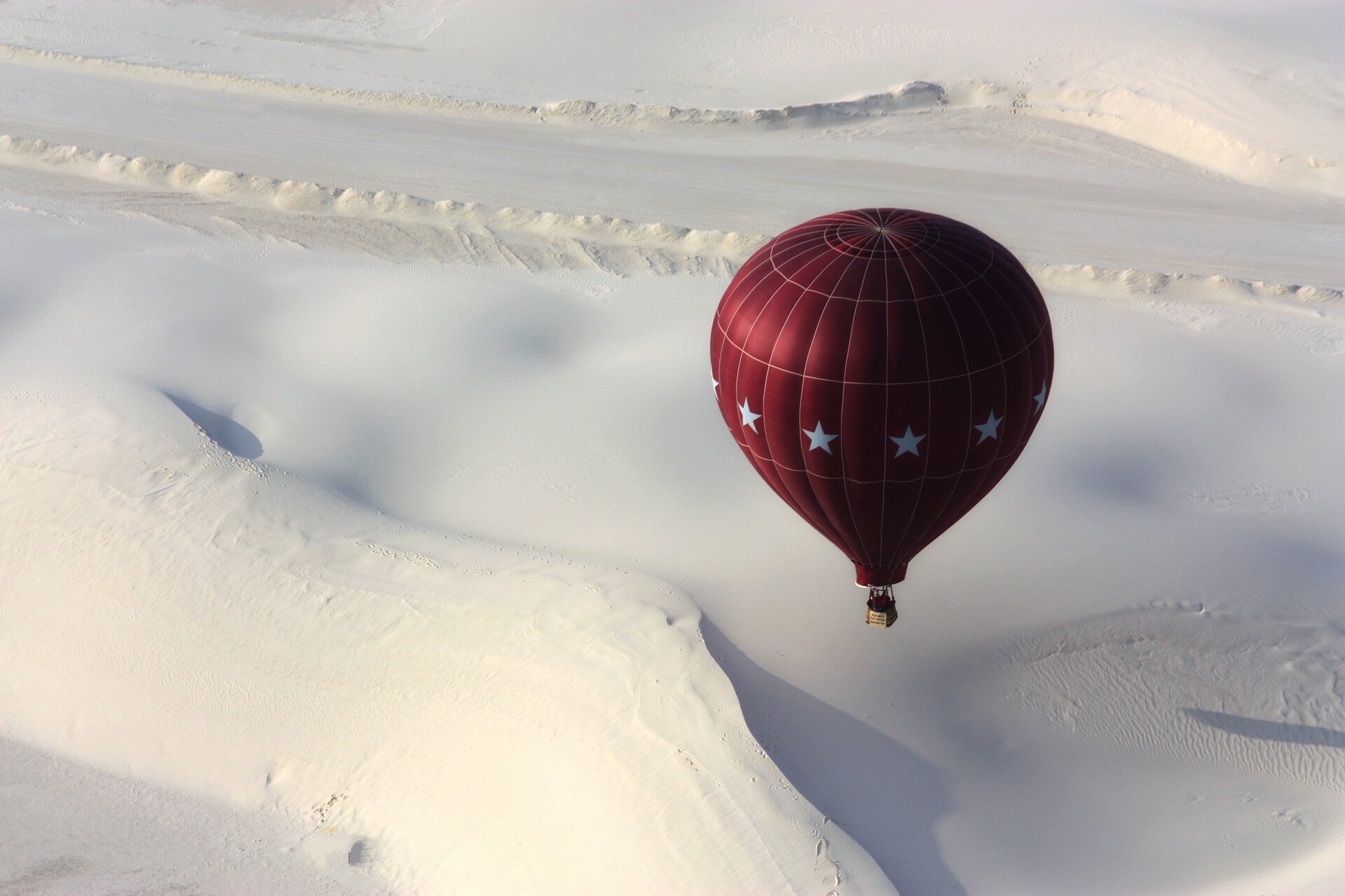 Zdjęcie lotnicze przedstawia park narodowy White Sands. Nad białą płaszczyzną zbudowaną z piasku leci czerwony balon z białymi gwiazdami z przymocowanym koszem.