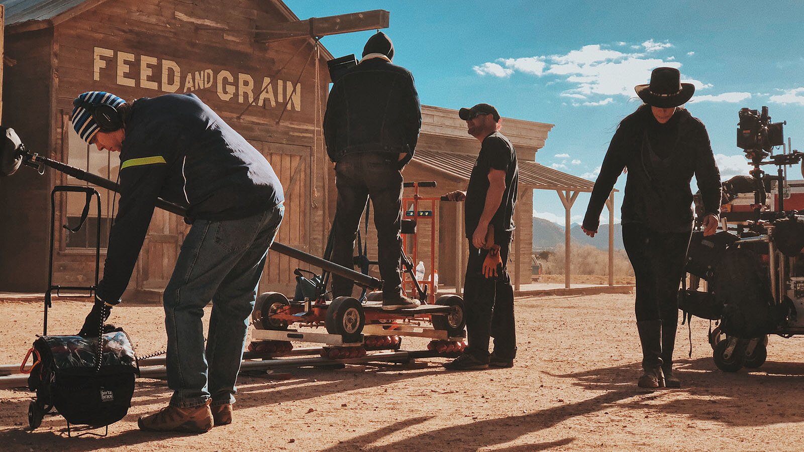 Zdjęcie pokazuje plan filmowy. Na pierwszym planie po lewej stronie są trzy osoby stojące obok wózka filmowego do kamery. Jedna osoba zakłada kamerę na wysięgnik, dwie stoją na i obok wózka przesuwającego wysięgnik. Po prawej stronie stoi mężczyzna i sprzęt filmowy. W tle znajduje drewniany budynek z napisem Feed and Grain.