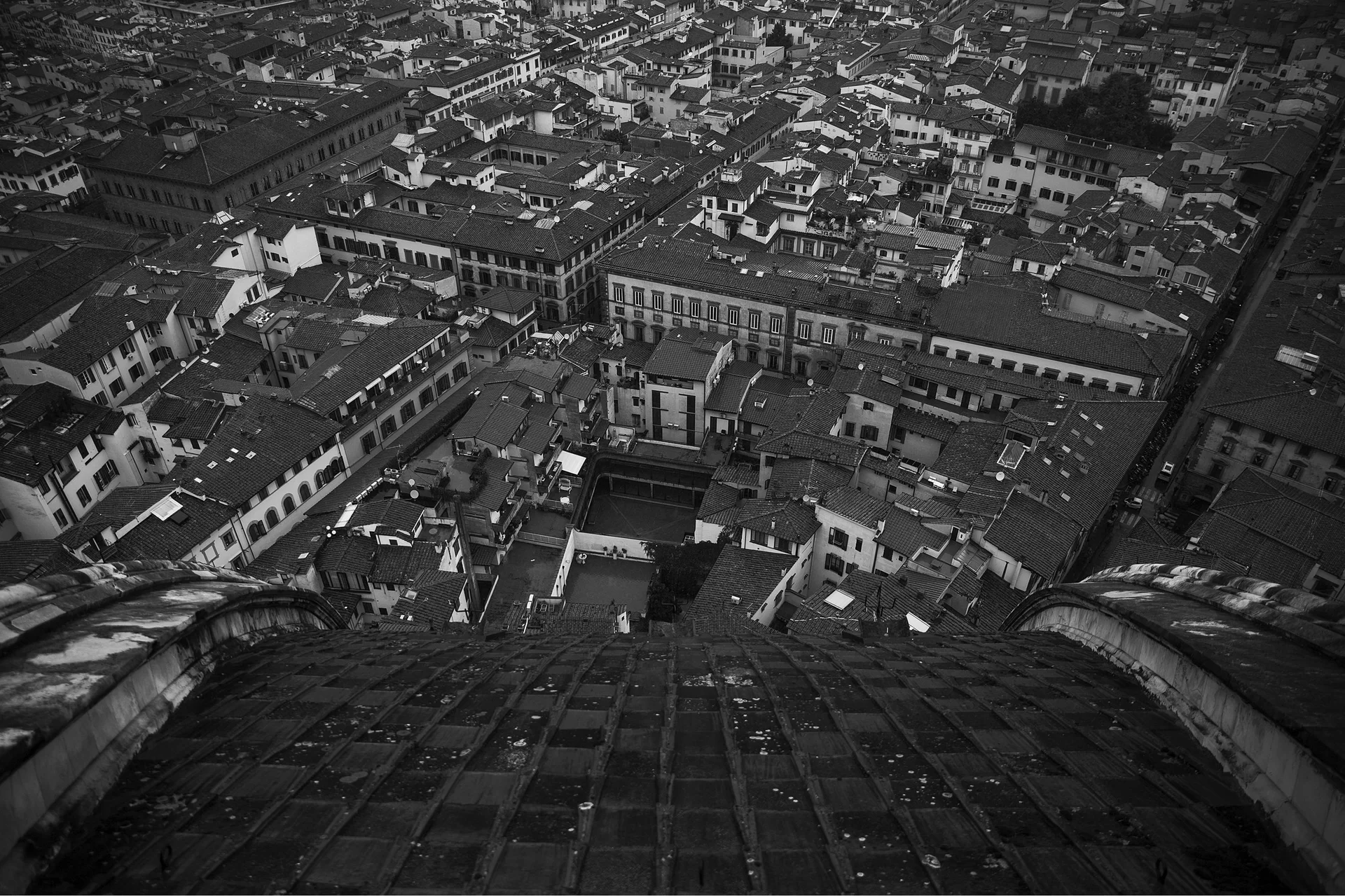 Ilustracja przedstawia zdjęcie Jakuba Wawrzaka. Czarno‑biała fotografia ukazuje widok na miasto ujęty z lotu ptaka. Pierwszy plan jest przybliżony i przedstawia fragment dachu. Budynki w tle zmniejszają się wraz z oddalaniem.