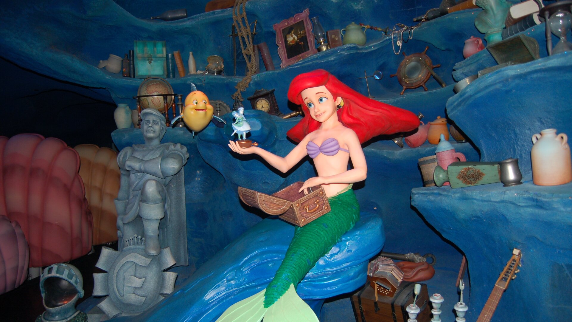 Kolorowa fotografia przedstawia aranżację komnaty Małej Syrenki z bajki Disneya. Syrenka Arielka ma czerwone włosy, fioletowy stanik i zielony ogon.  Siedzi na fali, na kolanach trzyma skrzynię ze skarbami, z której wyjmuje figurkę przedstawiającą tańczącego księcia i księżniczkę.  Po lewej stronie syrenki na wodnych półkach poukładane są różne przedmioty: mandolina, świecznik, kolejna skrzynia ze skarbami, klepsydra, wazon, naczynia. Po prawej stronie między zbroją rycerza a syrenką pływa żółta ryba.