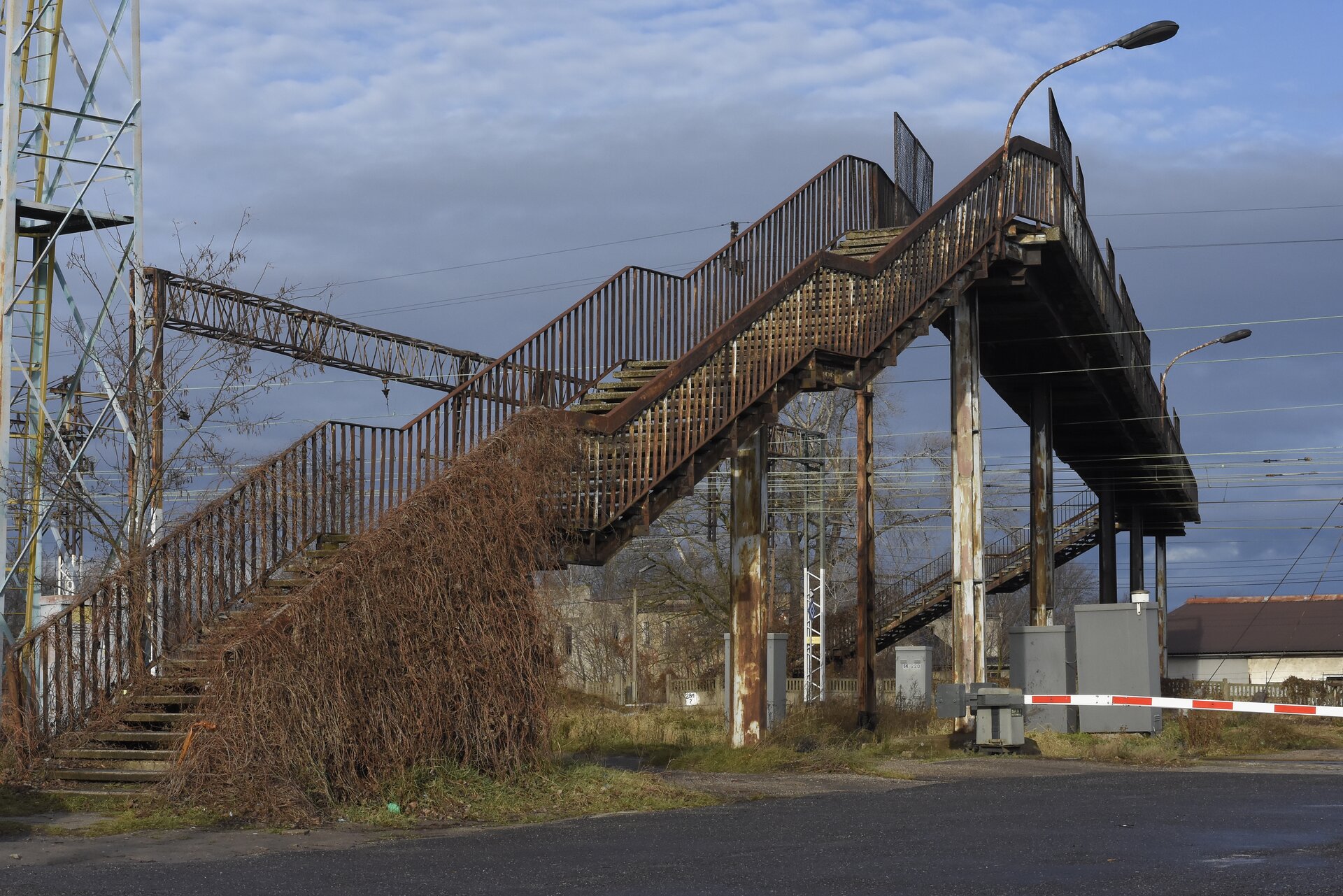  Ilustracja przedstawia kładkę dla pieszych, która biegnie nad torowiskiem. Aby skorzystać z kładki, należy skorzystać ze schodów. Barierka schodów jest pokryta uschniętą roślinnością. Na ilustracji widoczny jest także zamknięty szlaban kolejowy. Kładka wykonana jest z metalu, a w niektórych miejscach elementy kładki pokryte są rdzą. Nad kładką zamontowane są dwie lampy oświetlające przejazd kolejowy. Po lewej stronie ilustracji widać zarys przekaźnika, który jest wykonany z metalowej konstrukcji. Kładka umożliwia pieszym bezpieczne przekroczenie torów kolejowych.