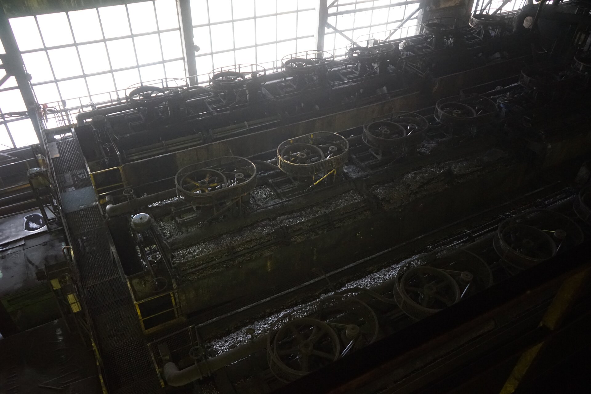 Na zdjęciu widoczny jest maszyna flotacyjna widoczna z góry. Maszna flotacyjna jest to urządzenie umożliwiające wzbogacanie fizykochemiczne za pomocą metody flotacyjnej. Na zdjęciu widoczna jest hala przemysłowa. W hali znajdują się maszyny flotacyjne. Maszyny flotacyjne tworzą trzy rzędy. Podstawą maszyny są podłużne prostopadłe stalowe pojemniki. Pojemniki są wypełnione czarną cieczą. Ciecz jest zbliżona konsystencją do szlamu. Pomiędzy pojemnikami utworzone są przejścia. Nad pojemnikami znajdują się stożkowe kształty. Na ich szczycie osadzone są okrągłe obiekty. Po obwodzie obiektów biegnie siatka. Sąsiadujące obiekty są połączone czarnym pasem.W tle zdjęcia widoczne są szyby.