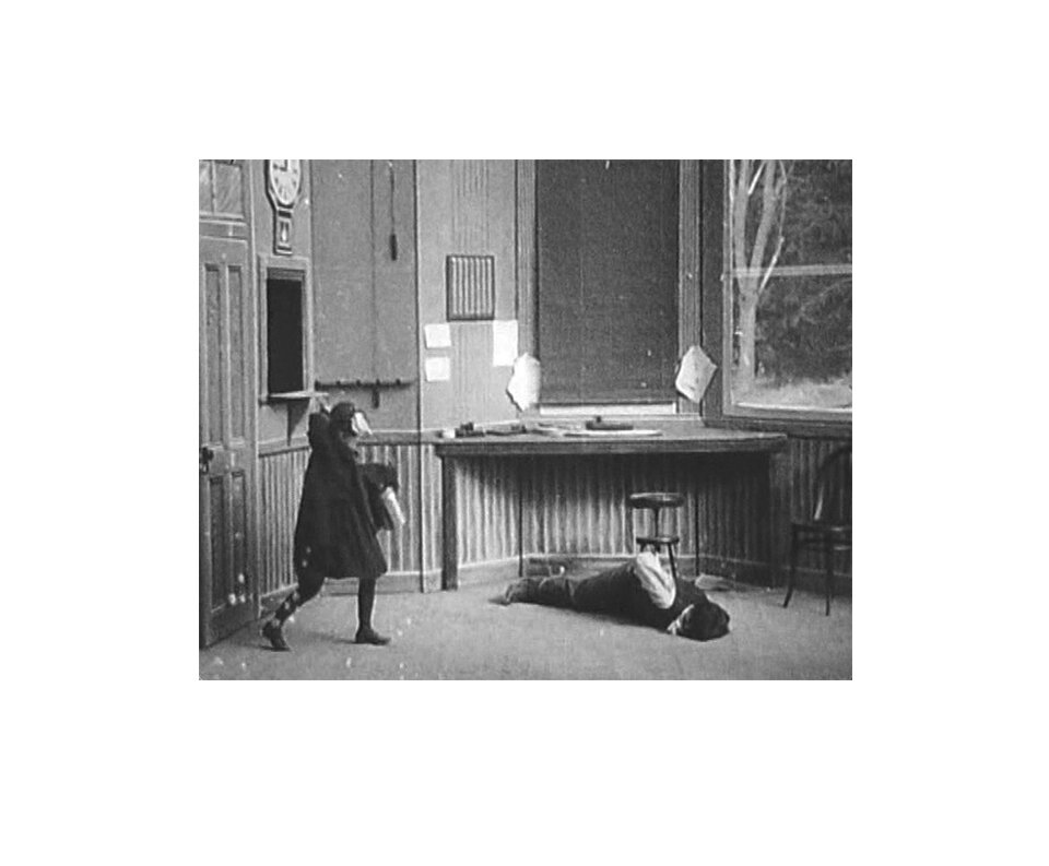 Kadr z czarno-białego filmu. Pomieszczenie to znane z pierwszego kadru wnętrze urzędu, prawdopodobnie dworca lub banku. Na środku kadru na podłodze leży mężczyzna w kamizelce, jest związany. Obok niego znajduje się dziecko, prawdopodobnie dziewczynka, w krótki płaszczyku. Dziewczynka podnosi rękę do góry w taki sposób, jakby chciała się zamachnąć na mężczyznę leżącego na ziemi. Dziecko trzyma w ręce jakiś przedmiot przypominający grubą książkę lub pudełko. Frame from black and white film. The room is the interior of the office, probably the train station or the bank, known from the first frame. In the middle of the frame on the floor lies a man in a vest, he is bound. Next to him is a child, probably a girl, in a short coat. The girl raises her hand up in such a way as if she wanted to swing at the man lying on the ground. The child holds an object in his hand resembling a thick book or box.